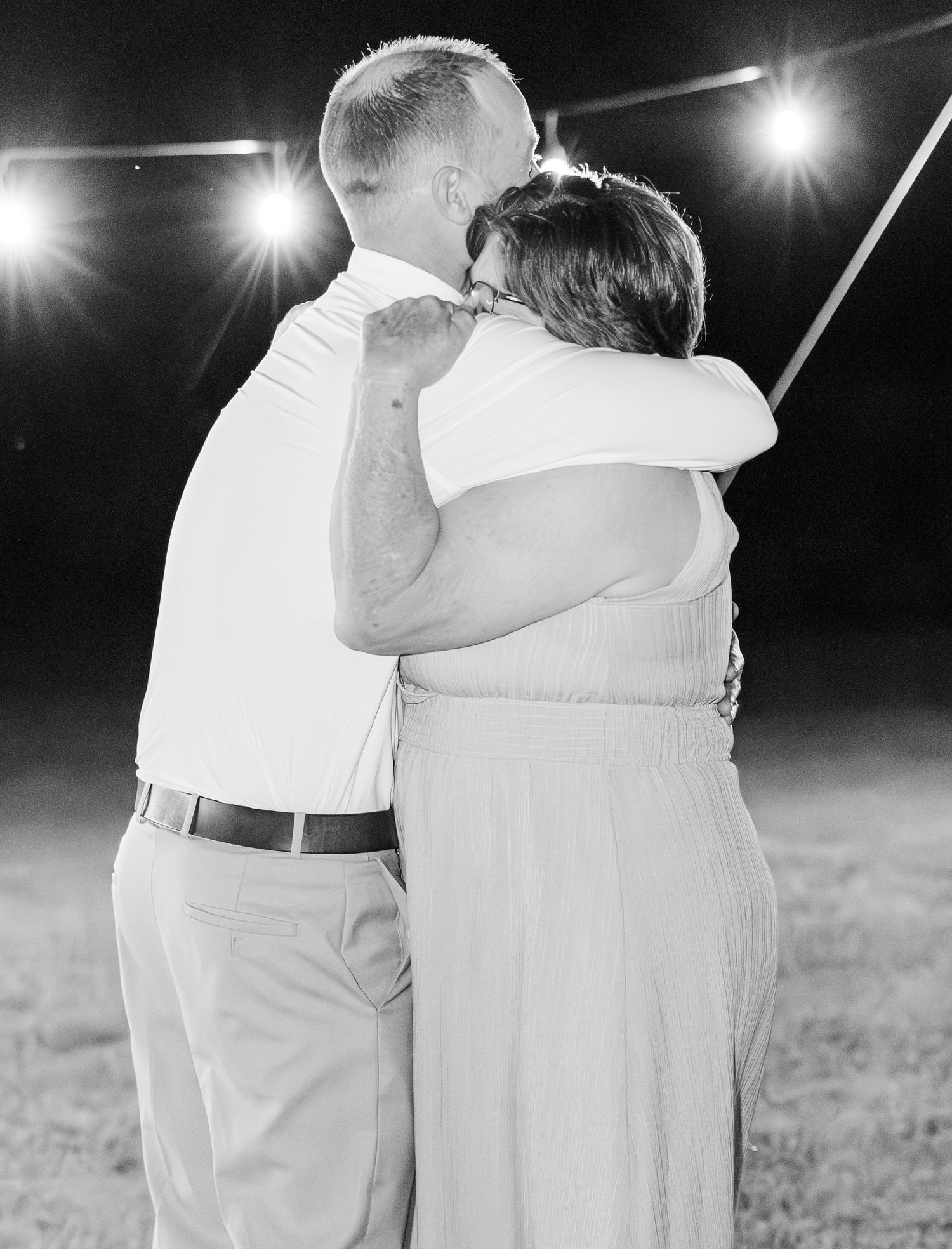 Black and white photo of groom dancing with mother under string lights at outdoor wedding reception.