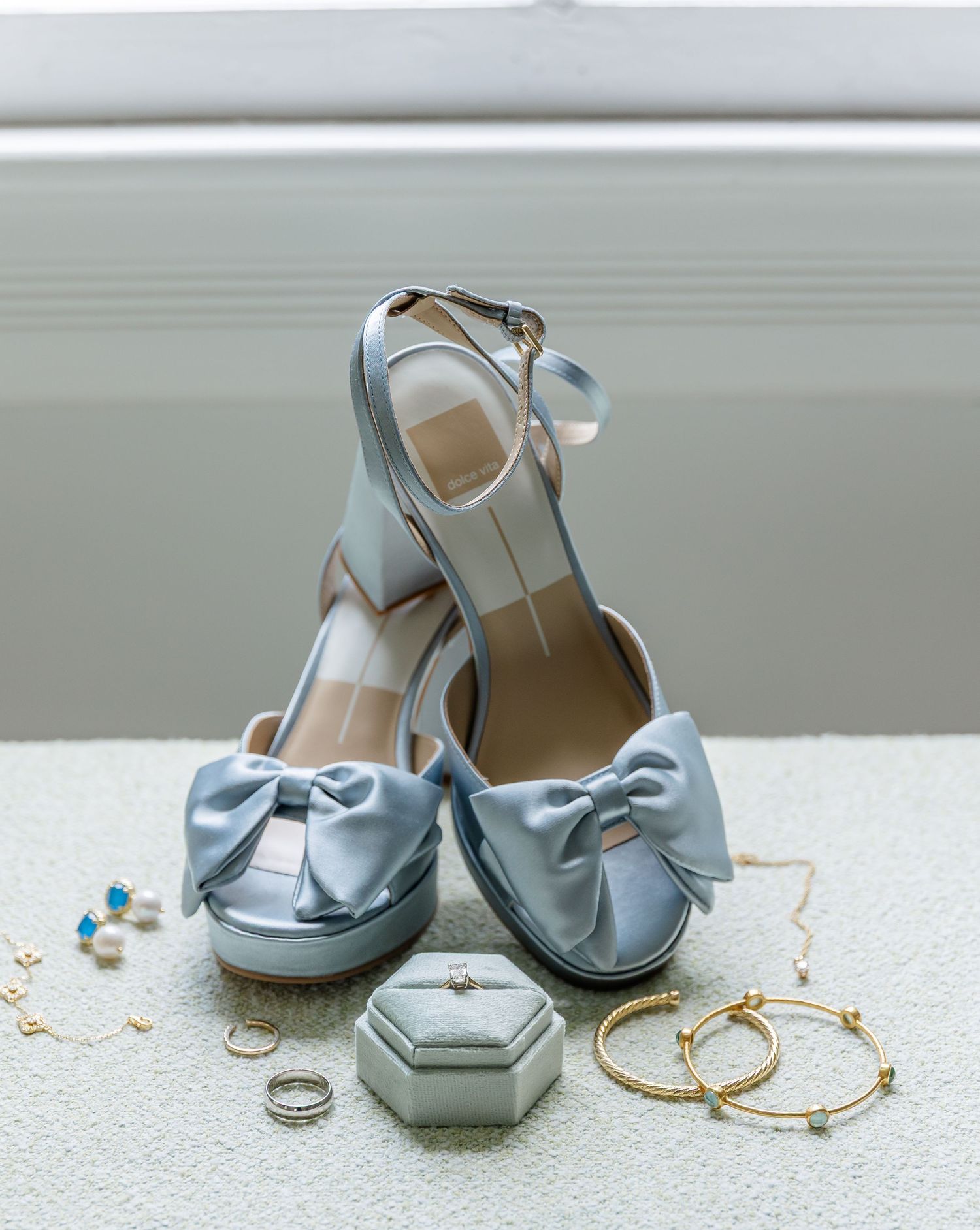 Pale blue wedding shoes with bows displayed with rings and jewelry on white surface.