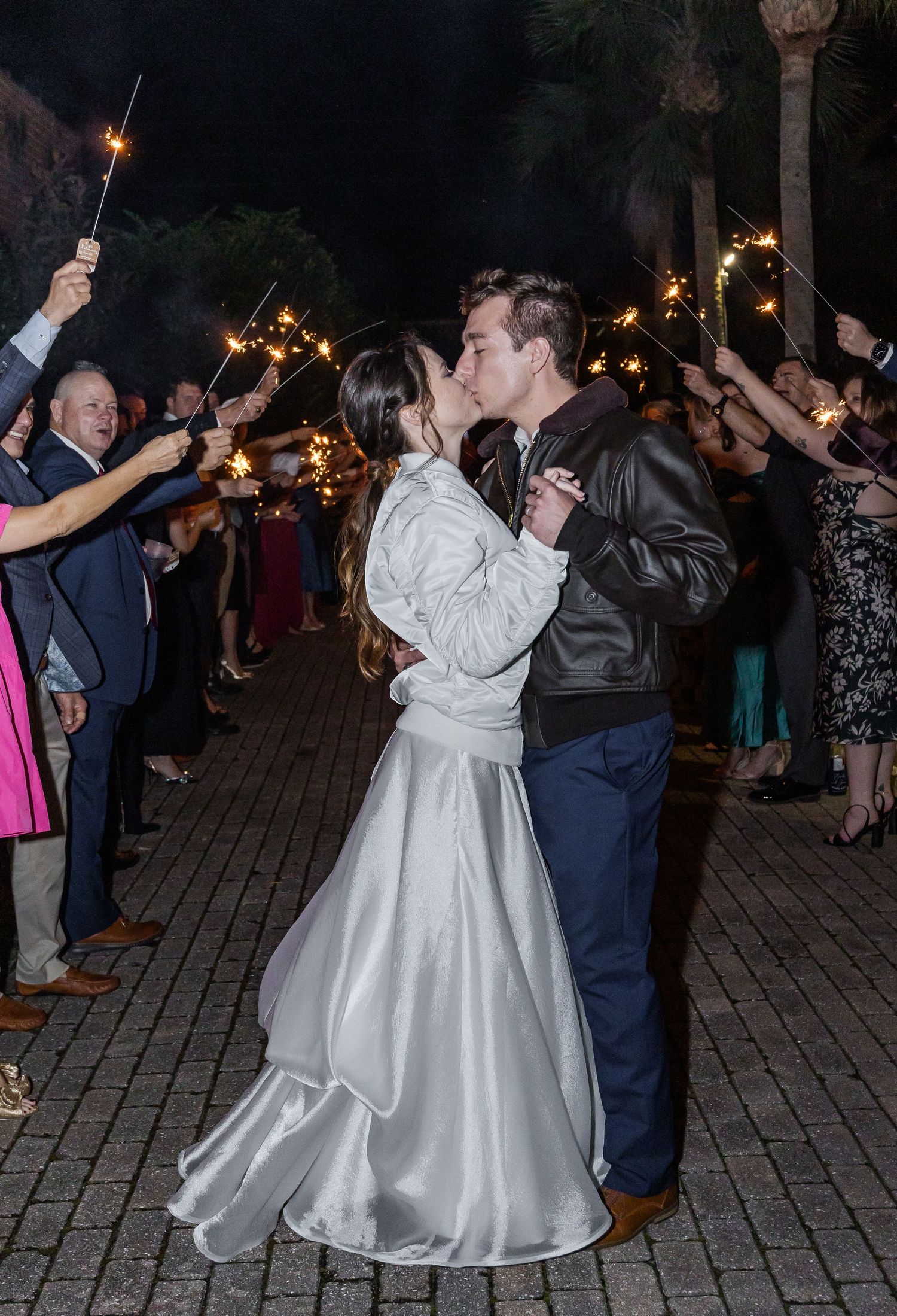 Wedding couple dances outside at night surrounded by guests holding sparklers.