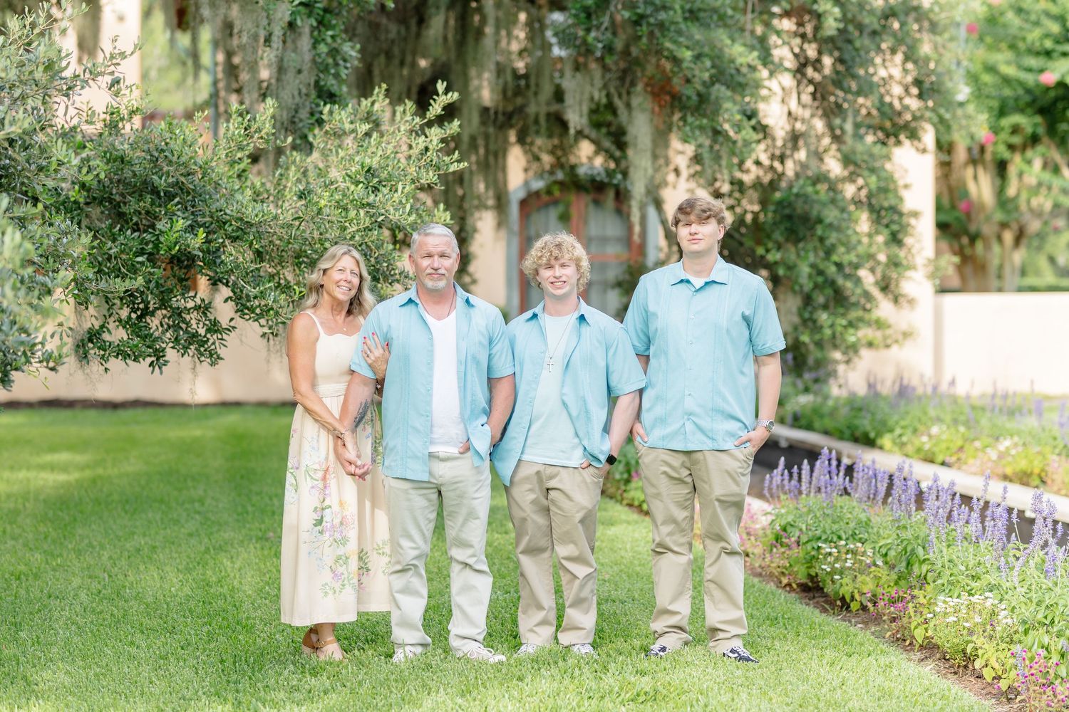 A group in coordinating light blue and cream outfits poses on a manicured lawn with trees and flowers in the background.