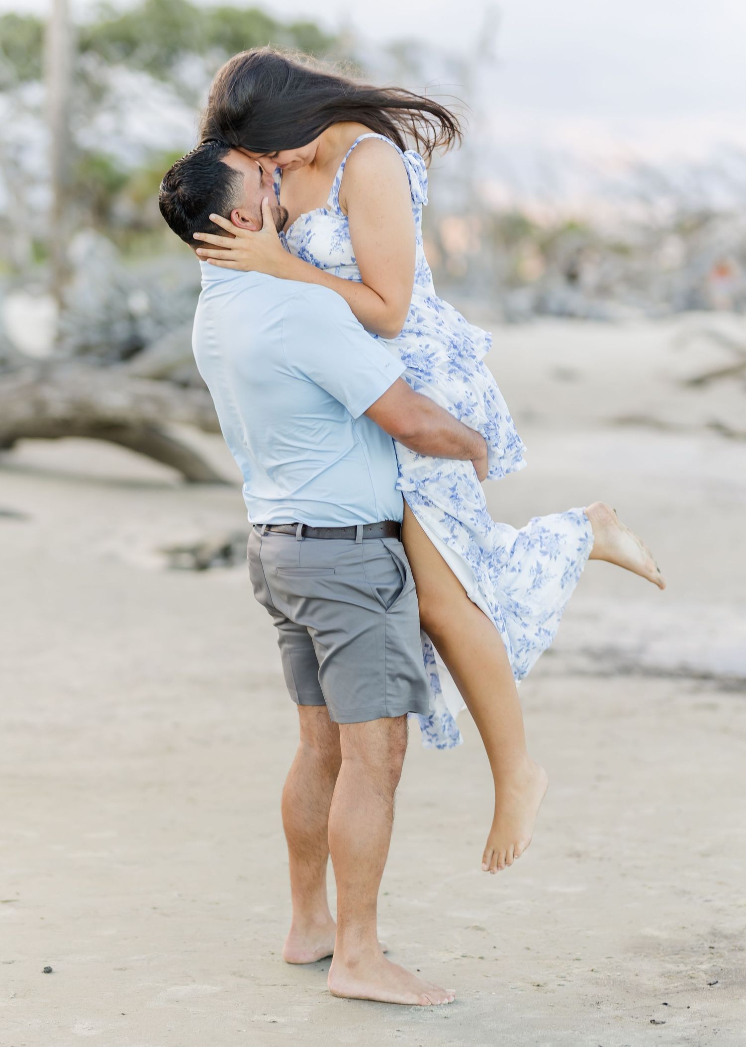 A romantic couple embraces on a sandy beach, with the person in blue lifting their partner wearing a floral dress.