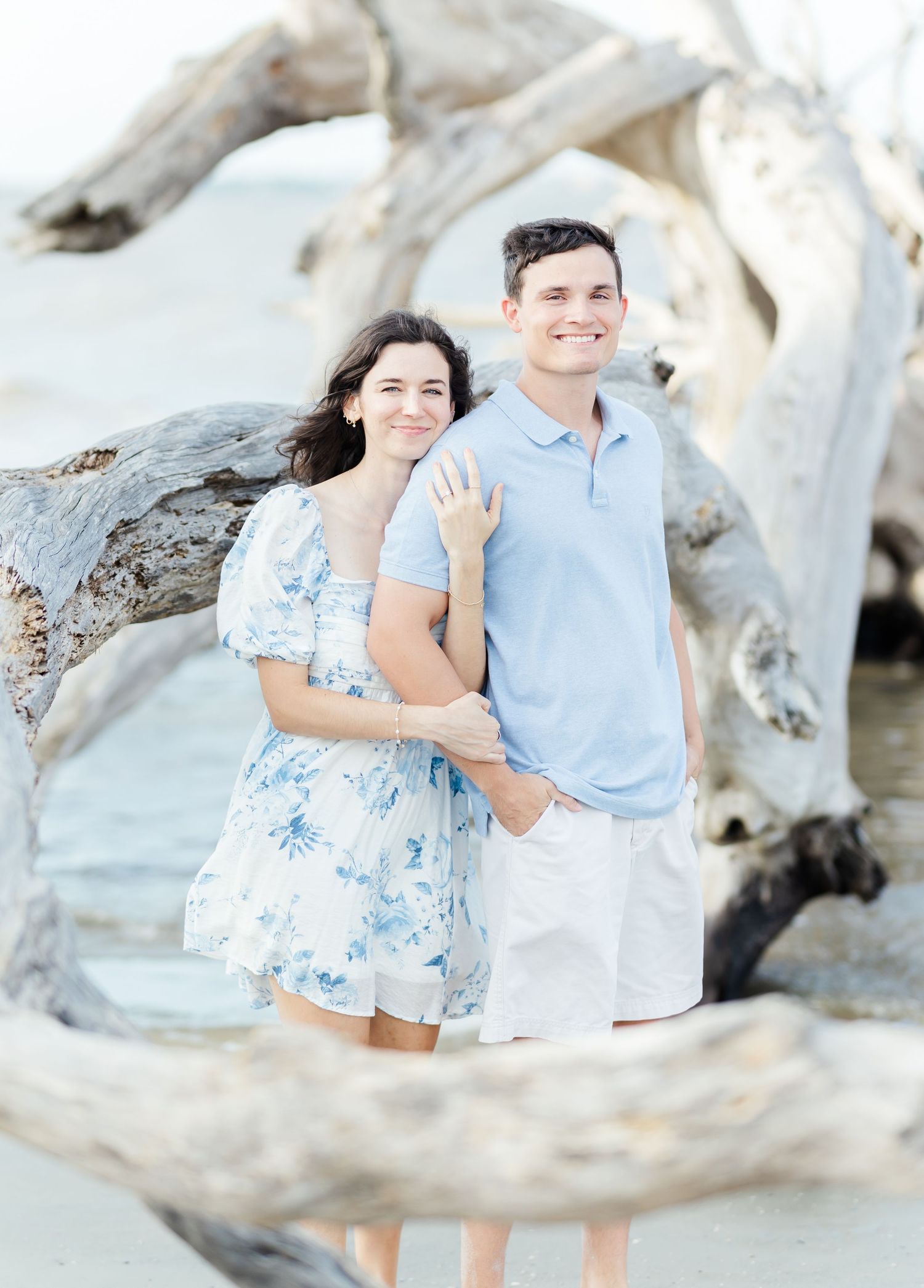 A romantic couple poses together on a driftwood beach while wearing light blue and white summer clothing.