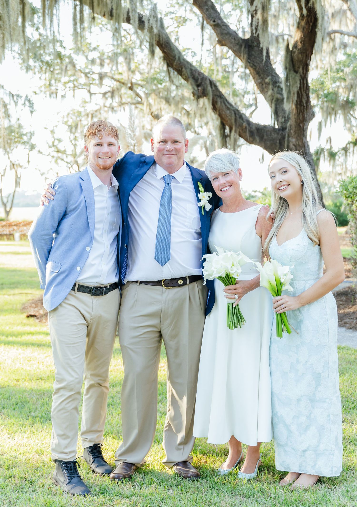 Wedding party in light blue and khaki attire poses together under Spanish moss-draped trees at outdoor venue.