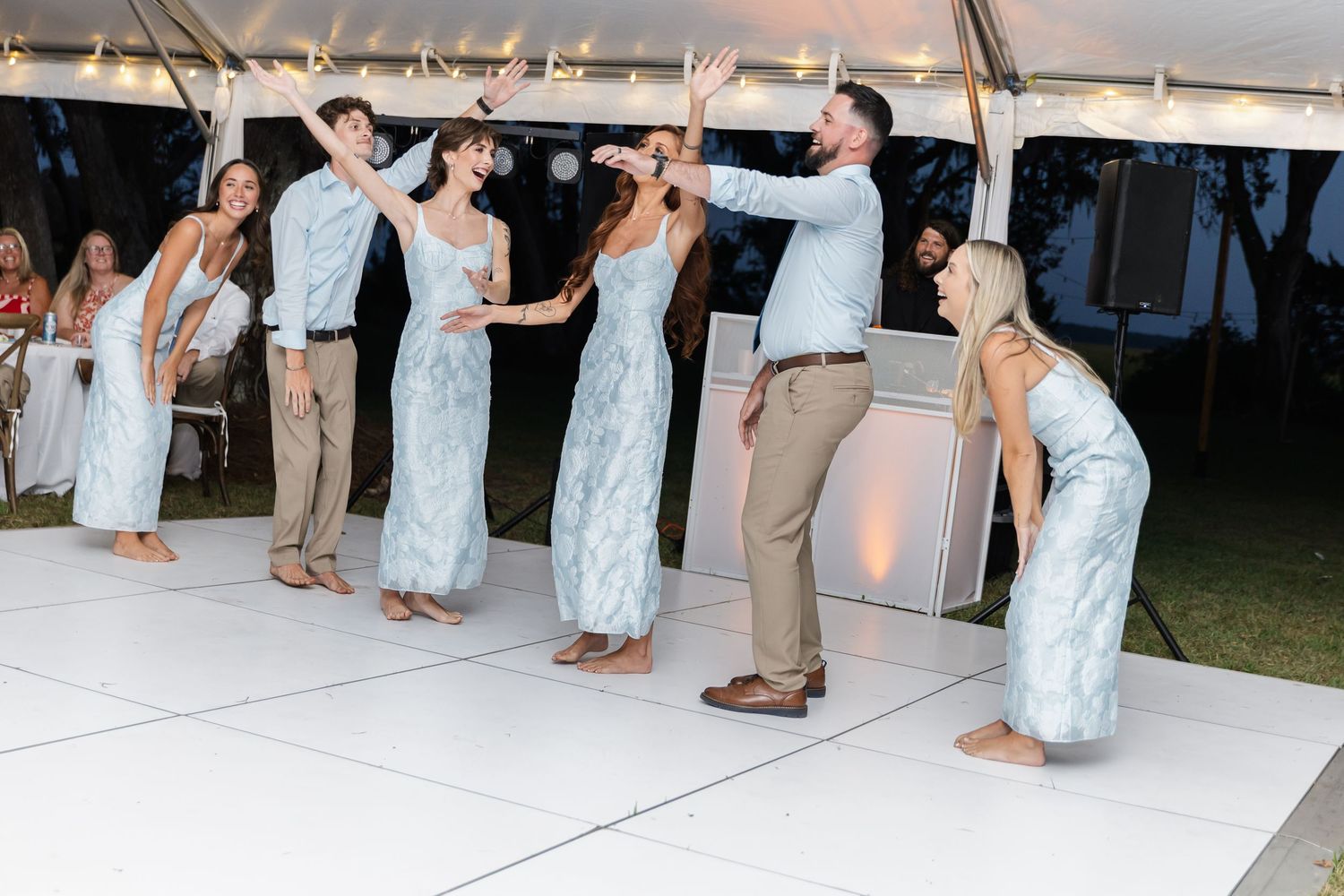 Wedding party dances on a white dance floor under a tent at an evening celebration.