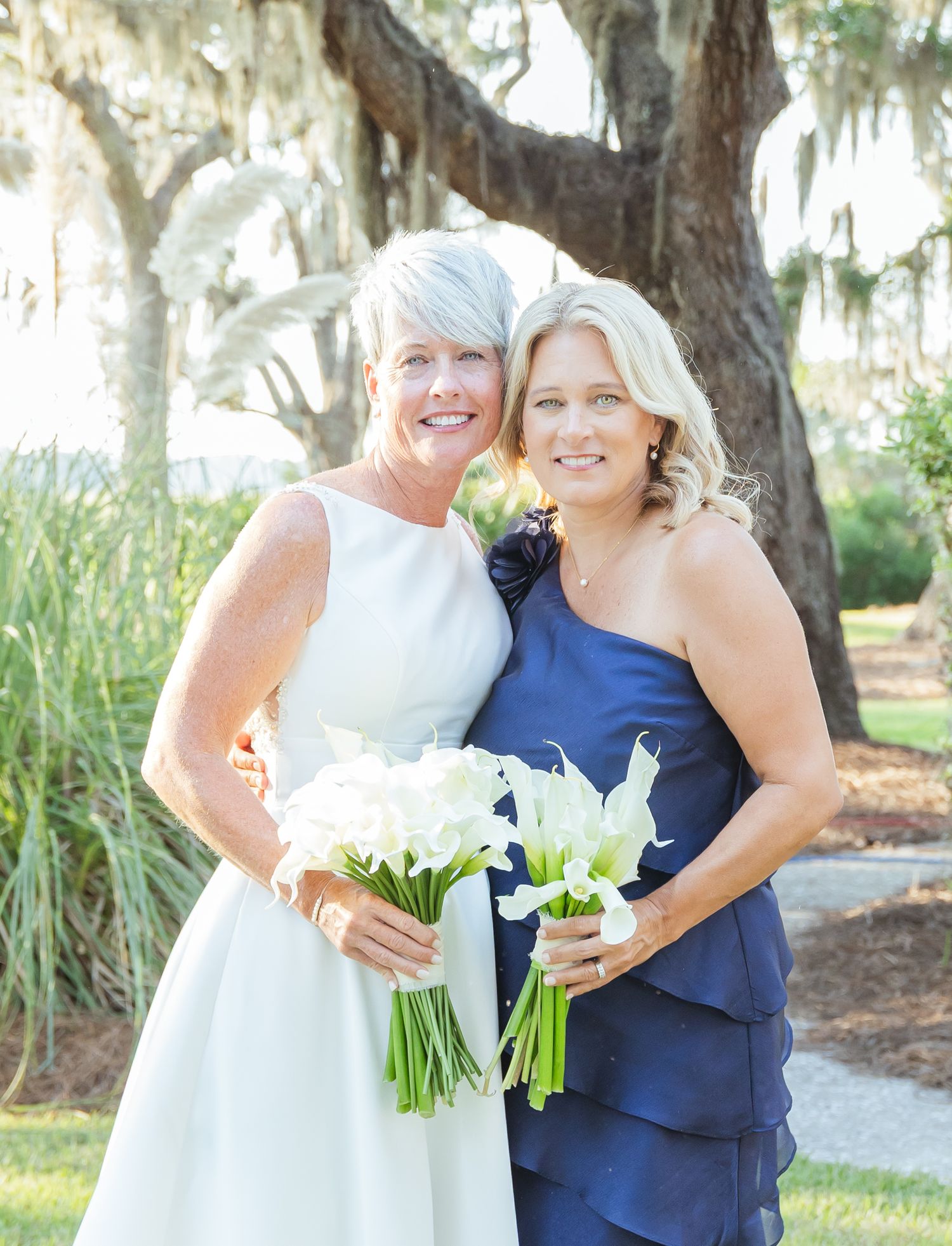 A group of people in navy blue and white dresses pose together outdoors under trees with bouquets.