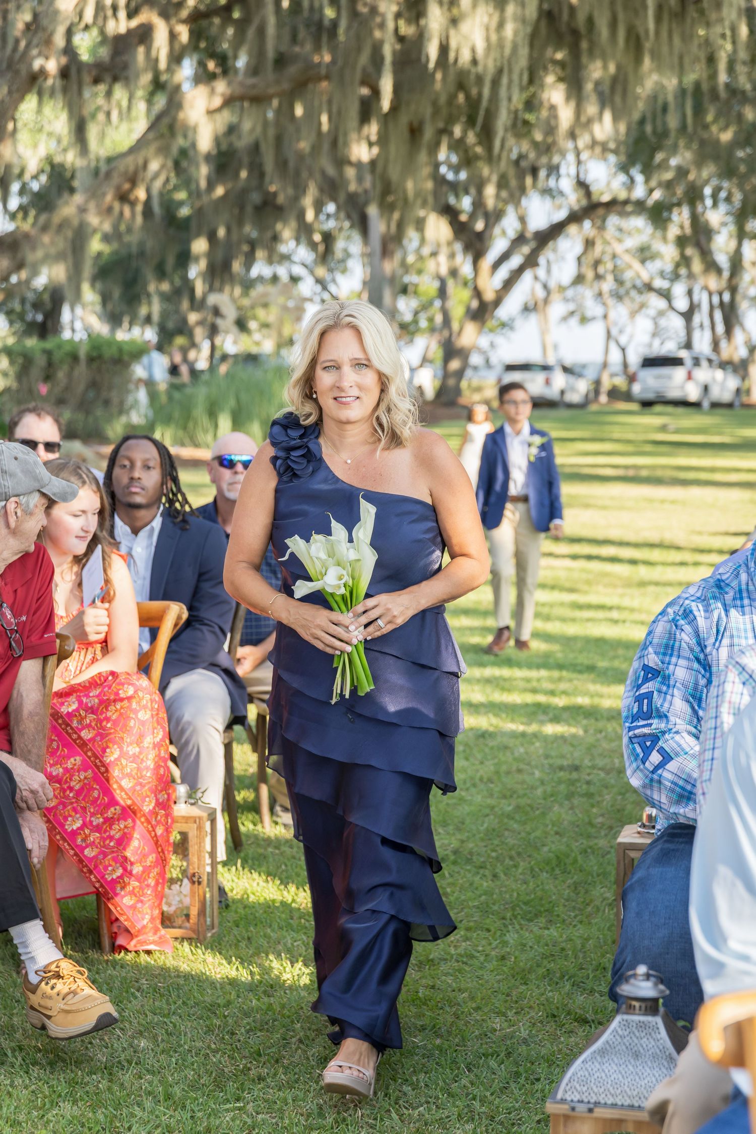 Wedding guests walk across a sunny lawn with Spanish moss hanging from trees in the background.