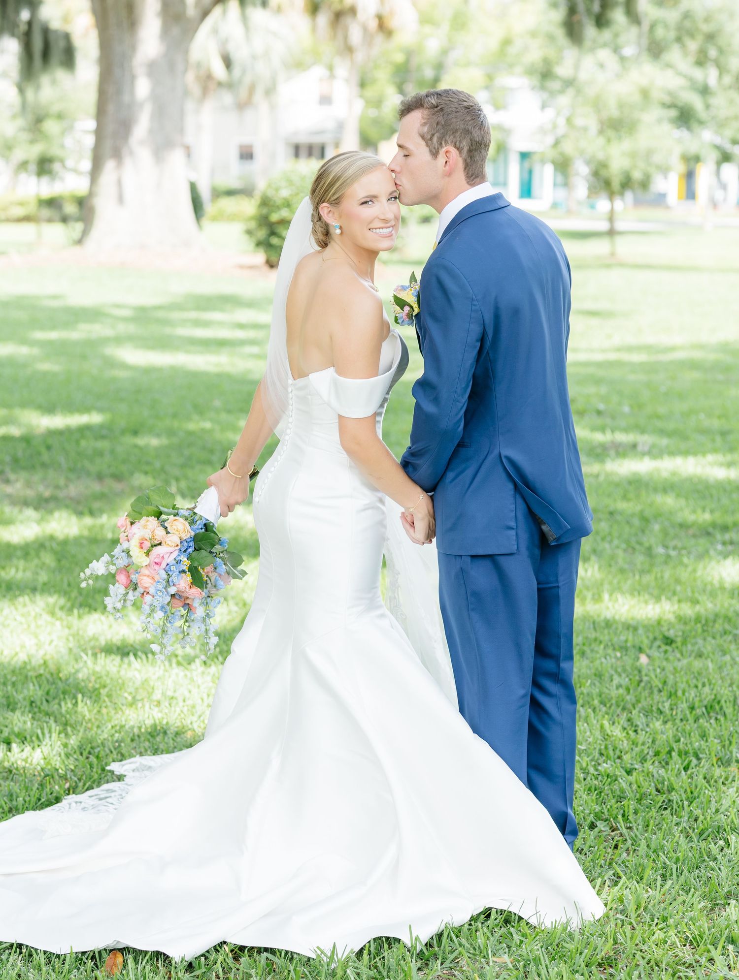 A couple in wedding attire shares a romantic moment on a sunlit lawn with pastel flowers and greenery.