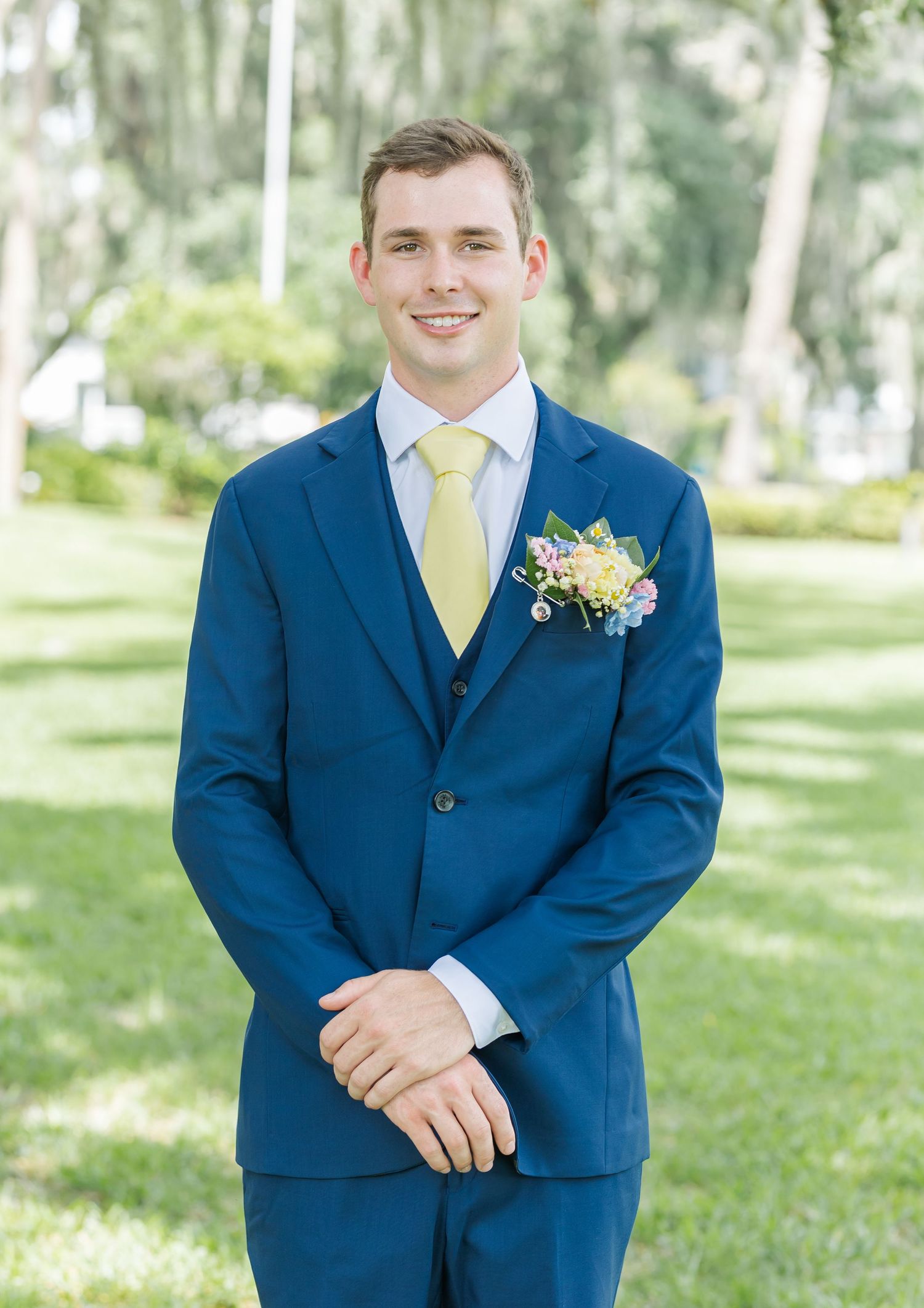 A groomsman in a navy blue suit with yellow tie poses outdoors in a garden setting.