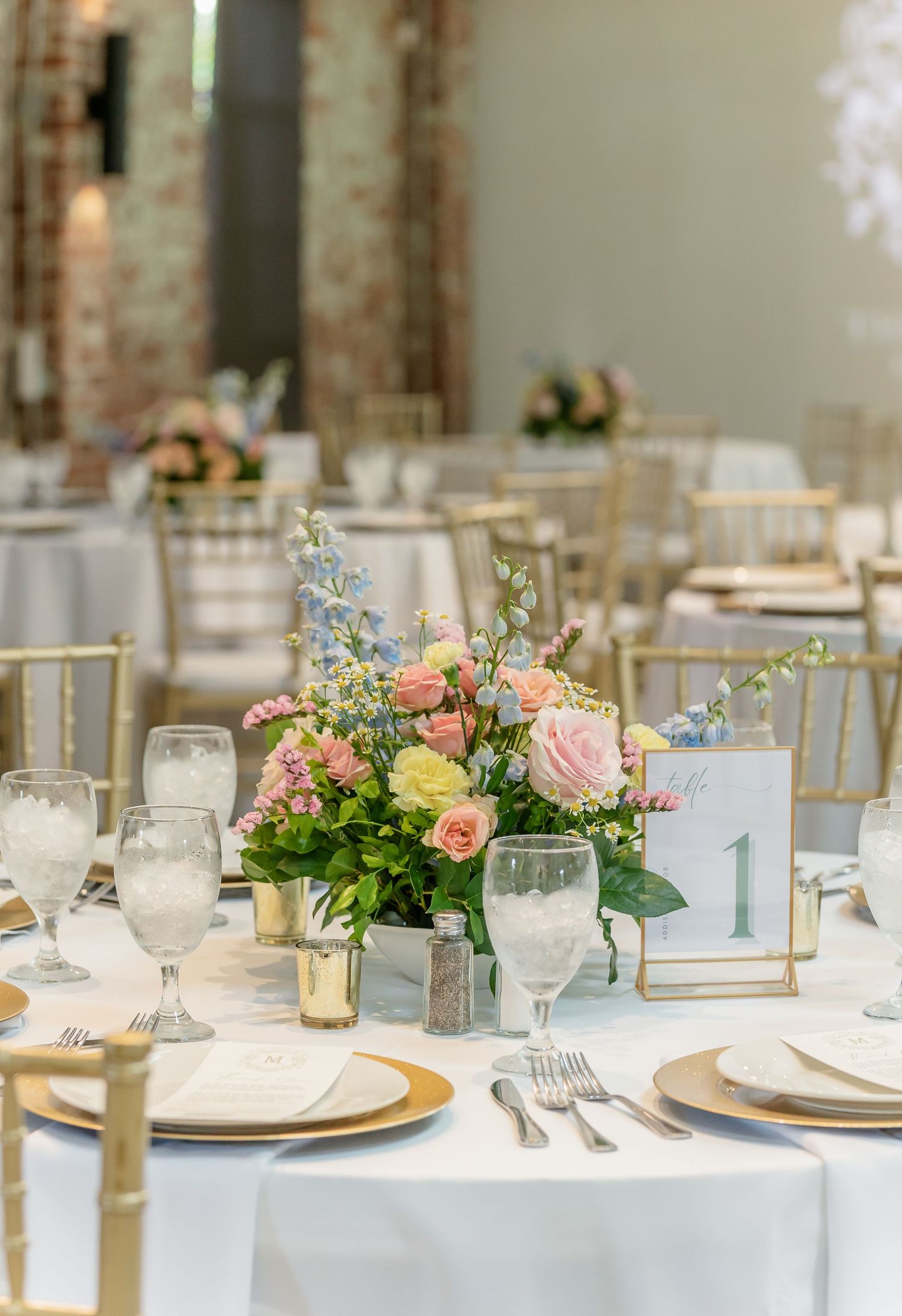 Elegant wedding reception table setting with white linens, gold accents, and soft pink and yellow floral centerpieces.