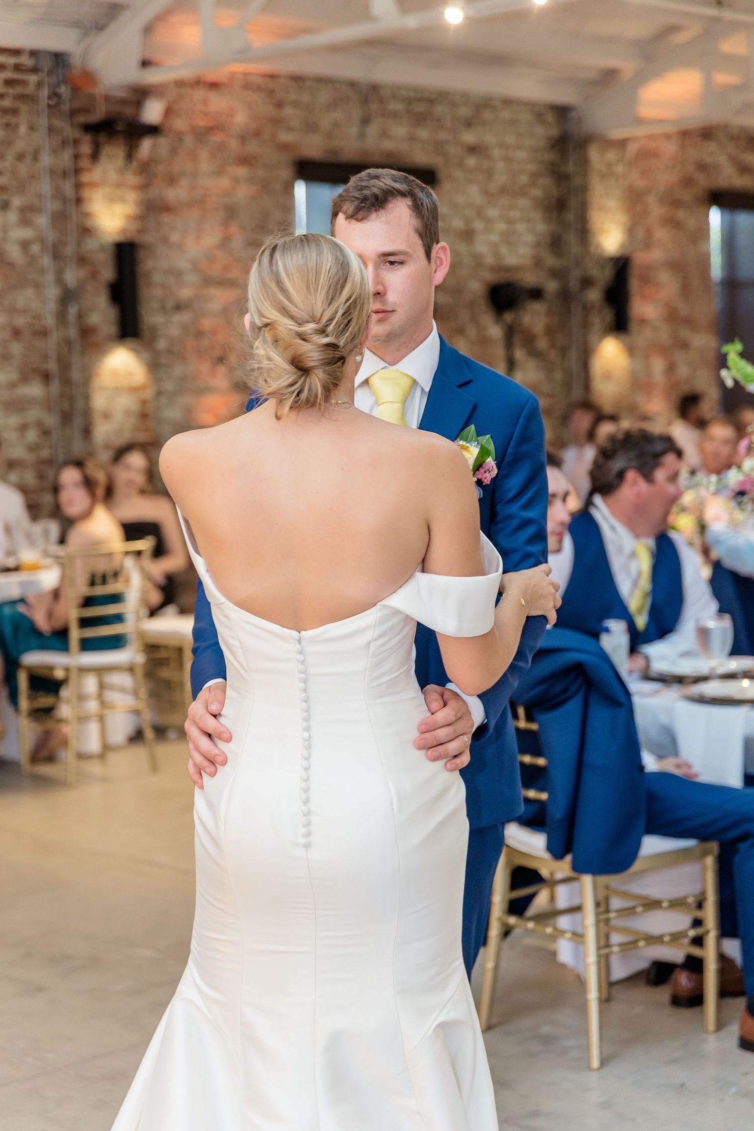 A bride and groom share their first dance at a rustic brick venue with elegant lighting and wedding guests looking on.
