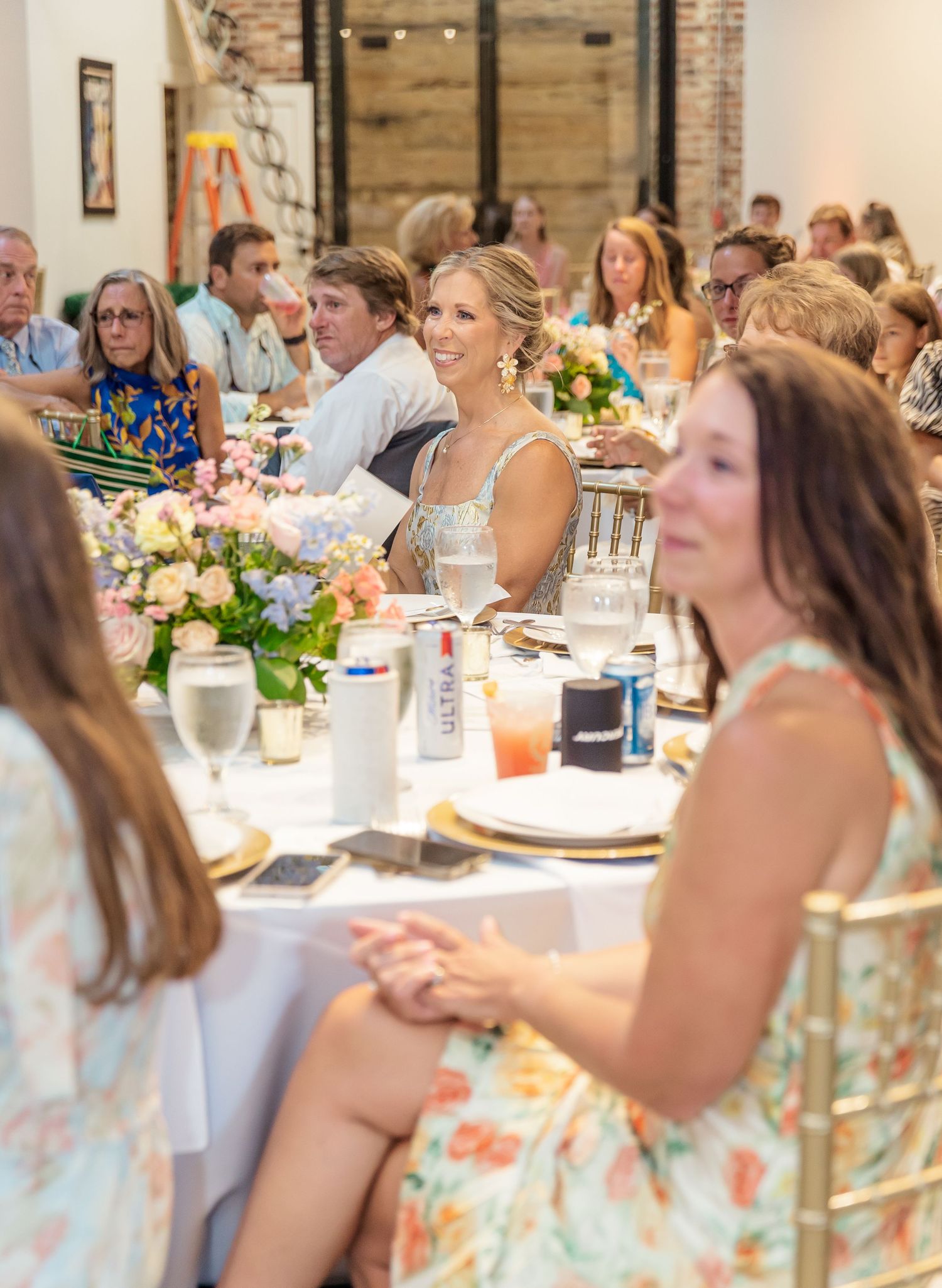 Wedding guests enjoy a seated dinner reception in a rustic brick venue with elegant table settings and floral centerpieces.