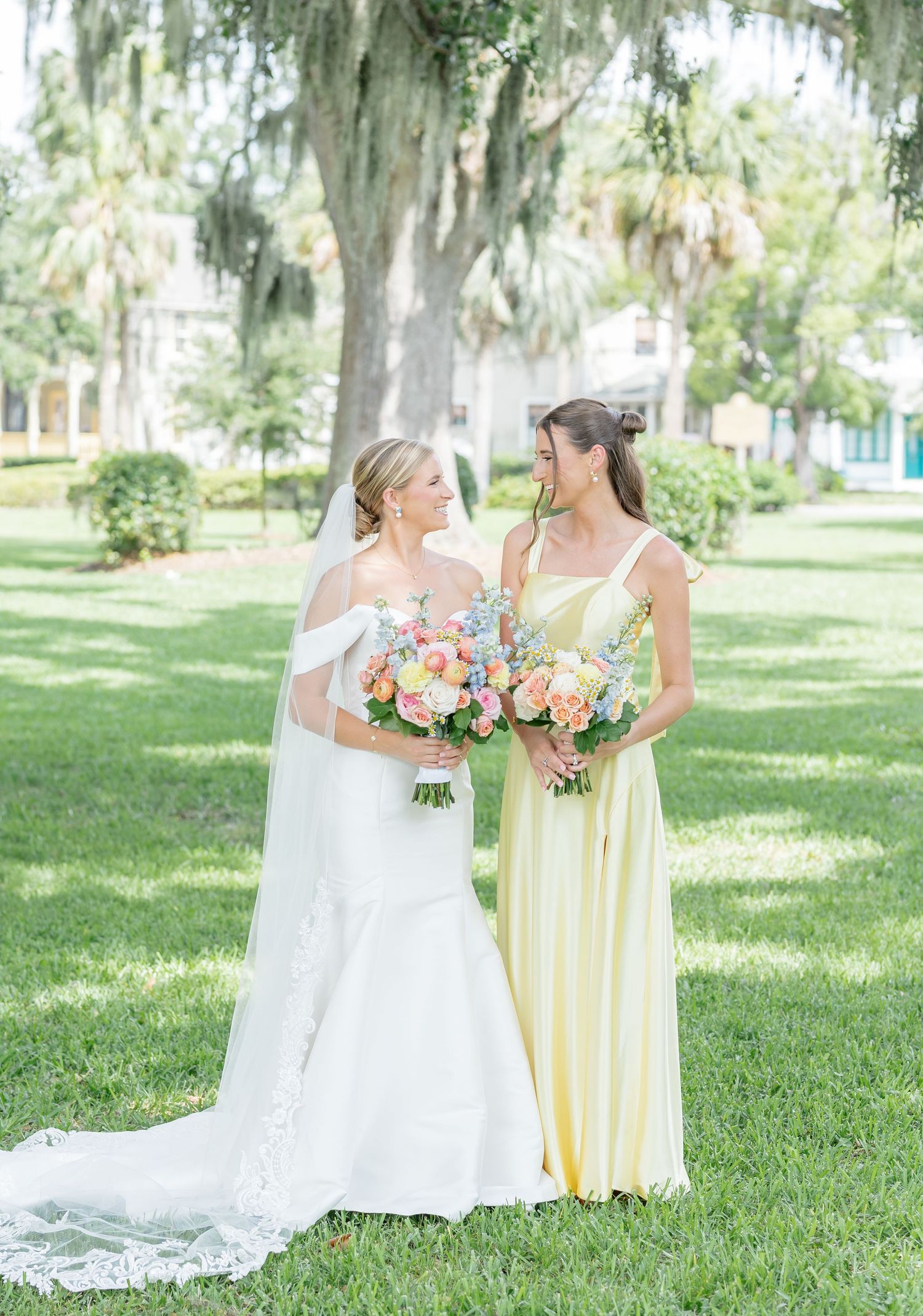 Two people in yellow and white dresses stand together holding pink floral bouquets in a sunny garden setting.