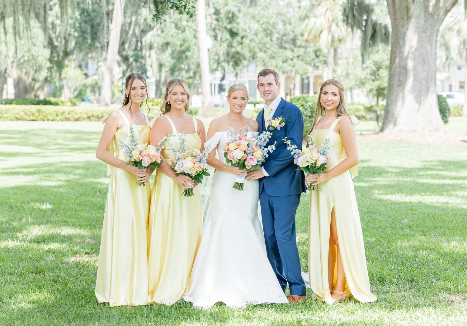 Wedding party in yellow bridesmaid dresses and navy suit poses together on sunny lawn under oak trees.