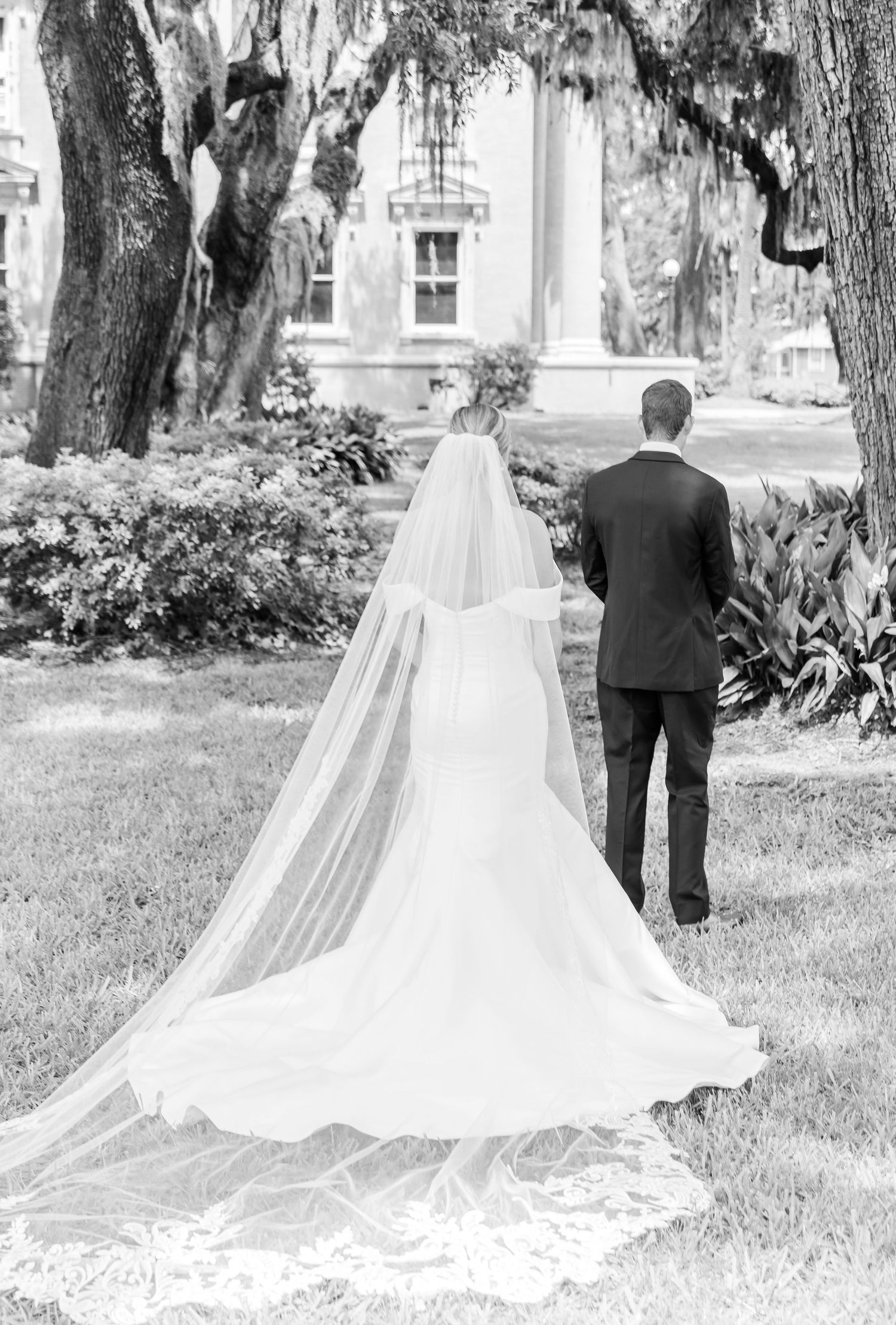A black and white wedding photo showing a bride in a flowing cathedral veil and gown standing with groom under oak trees.
