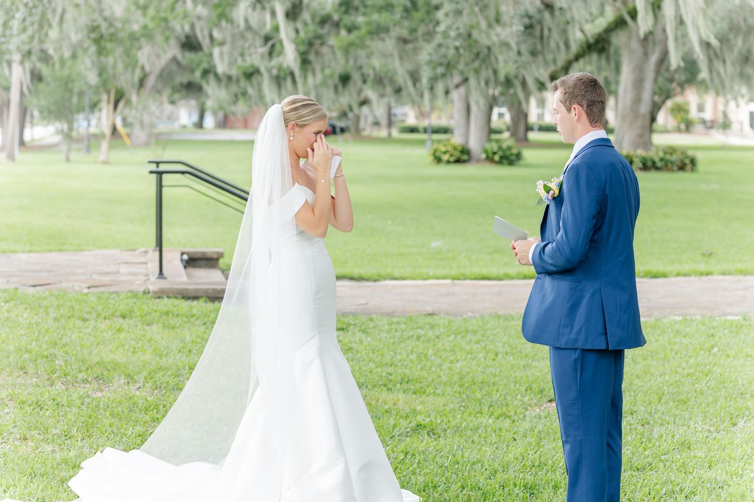 A couple in formal wedding attire shares a romantic moment in a lush green outdoor garden setting.