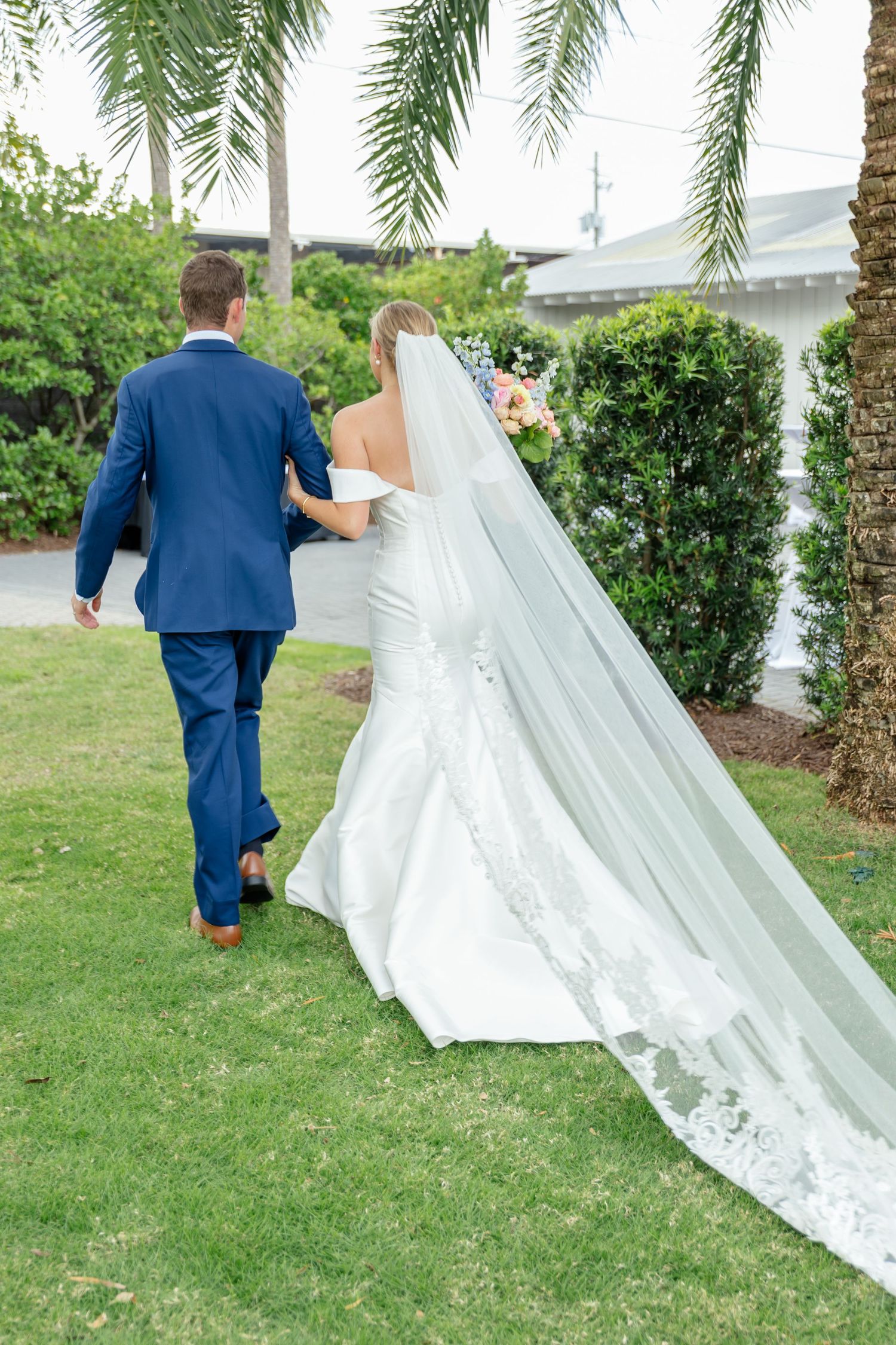 A couple walks on green lawn with a flowing cathedral-length lace veil trailing behind in an outdoor garden setting.