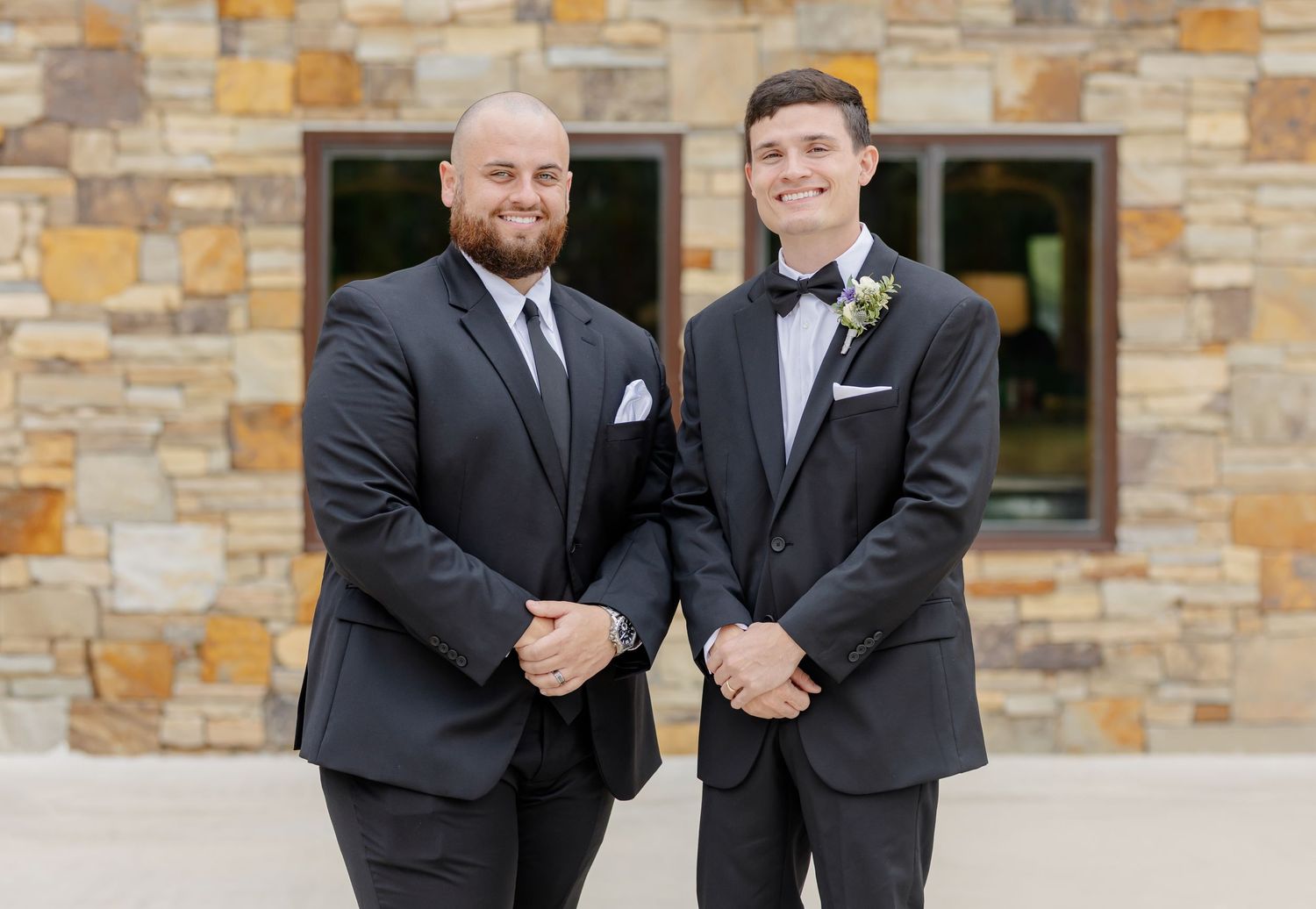 Two wedding party members in matching black suits share a candid moment against a stone facade.