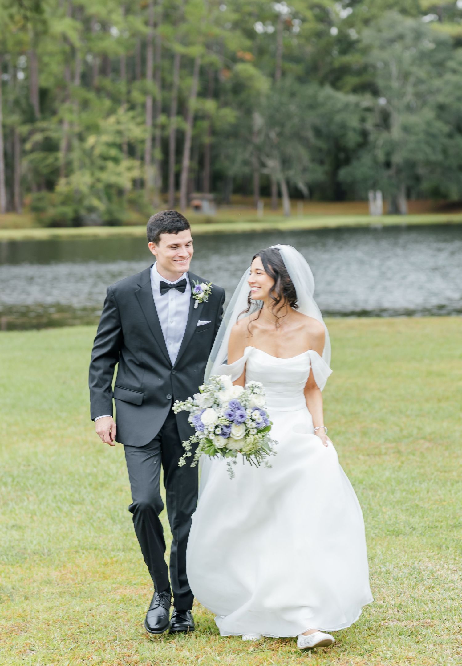 Playful wedding couple runs through grassy field near lake with flowing white dress and bouquet.