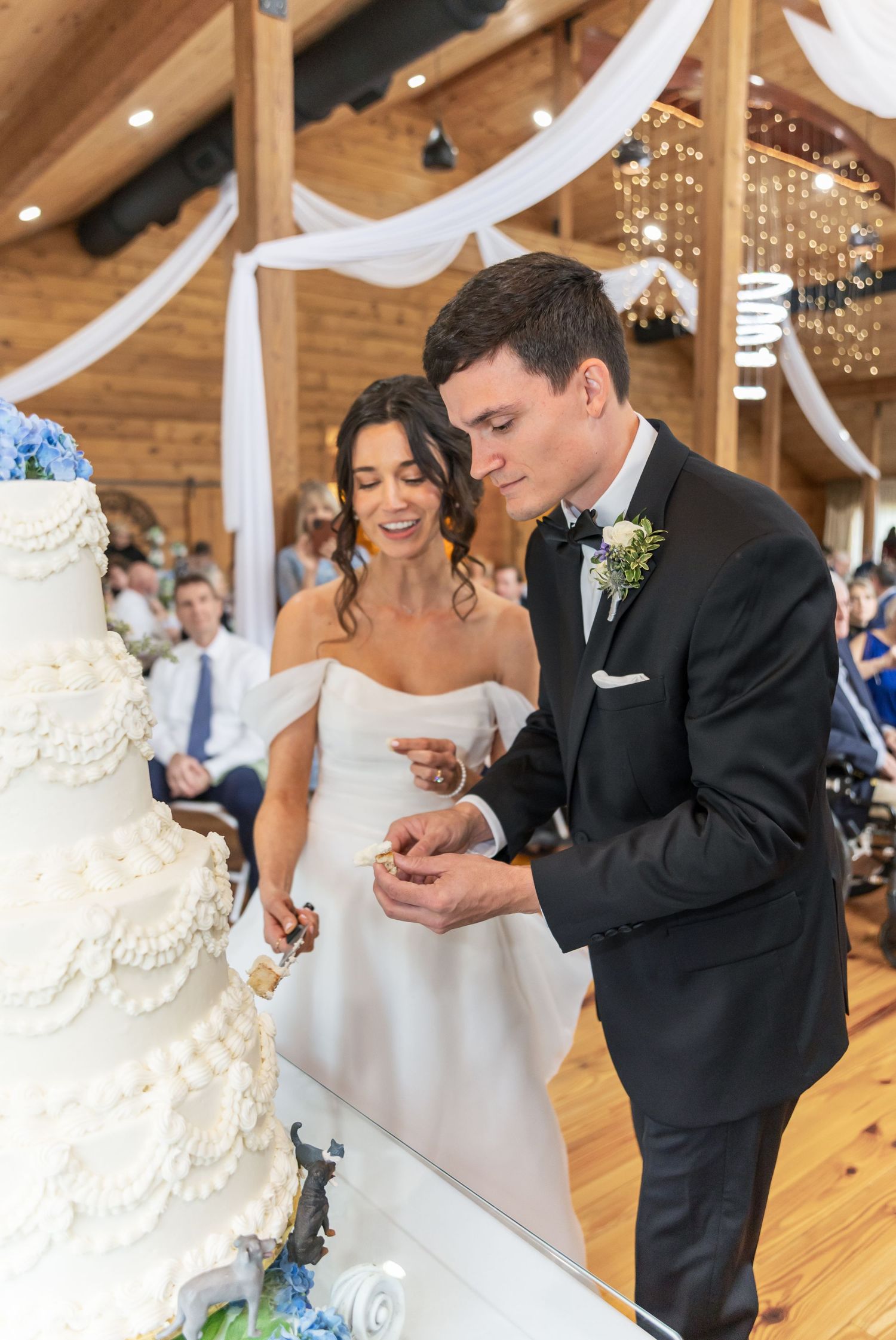 Wedding couple shares a sweet moment cutting their elegant white tiered cake at an indoor rustic venue.