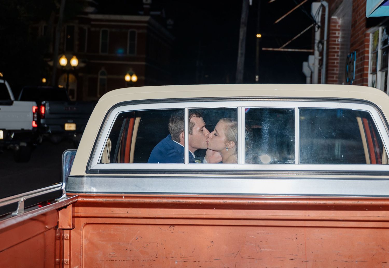 Two people kiss in a classic white car parked at night with illuminated street lamps visible in the background.