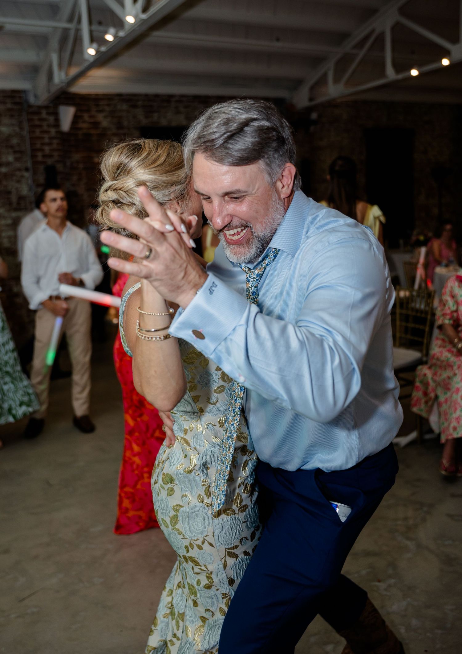 A couple dances together at an indoor venue, with the man in a light blue shirt and the woman in a patterned dress.