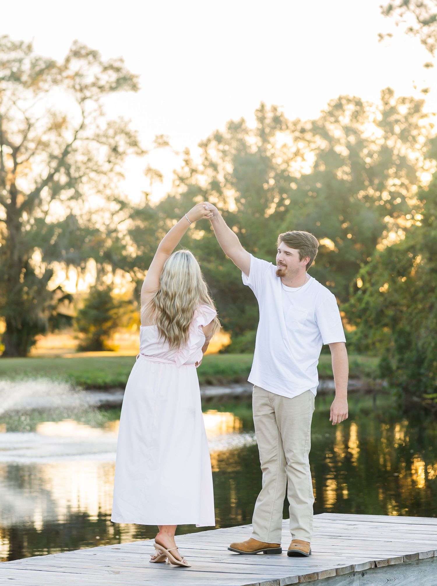 Dancing couple twirls on wooden dock with water fountain and sunset in background.