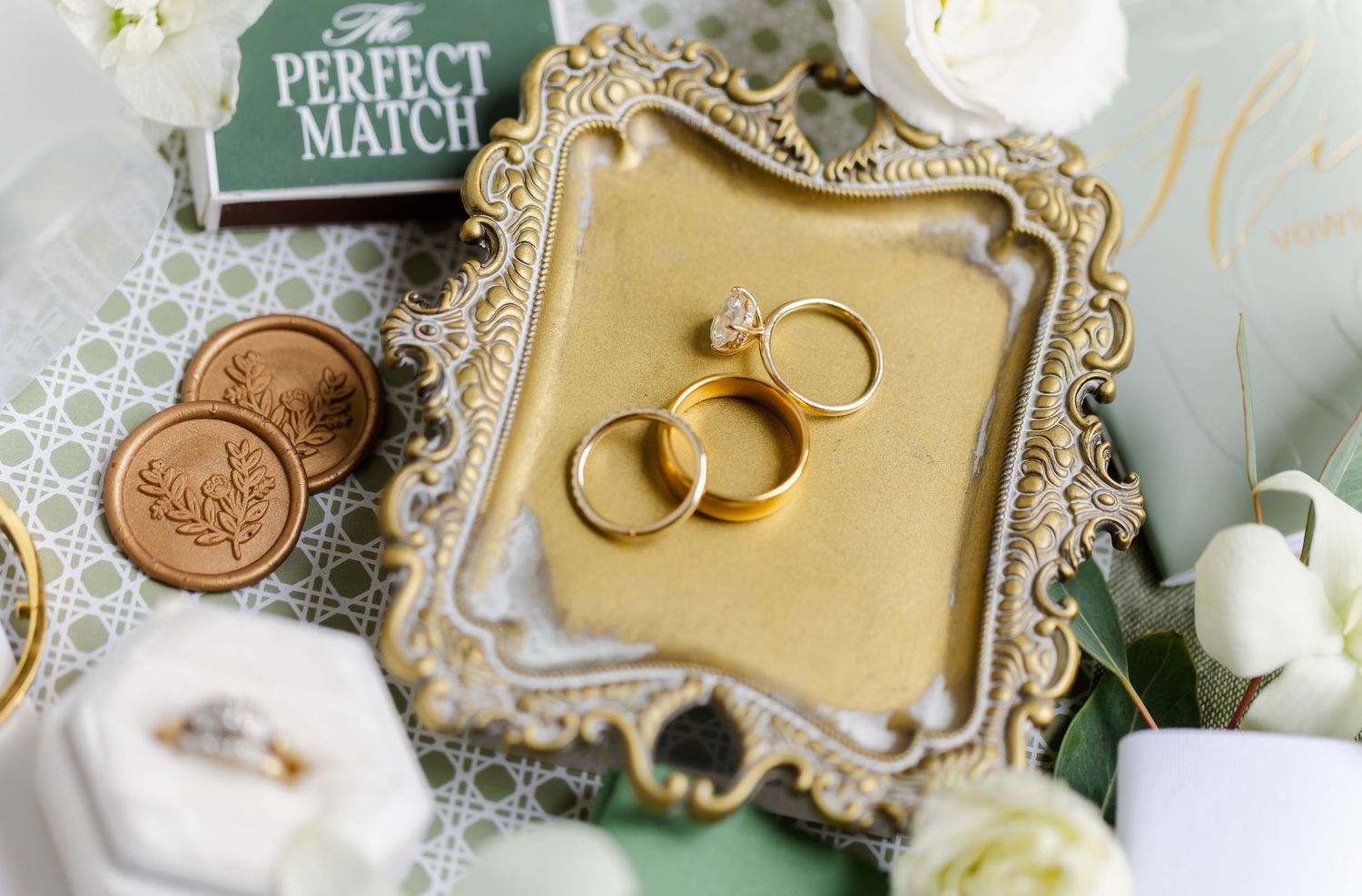 Wedding rings and pearl jewelry displayed on ornate gold frames with delicate white floral accents.