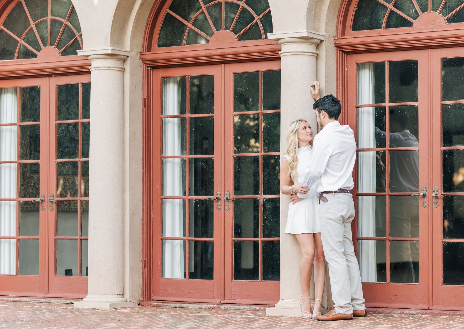 A romantic couple embraces in front of elegant coral-colored French doors with arched windows at a historic venue.
