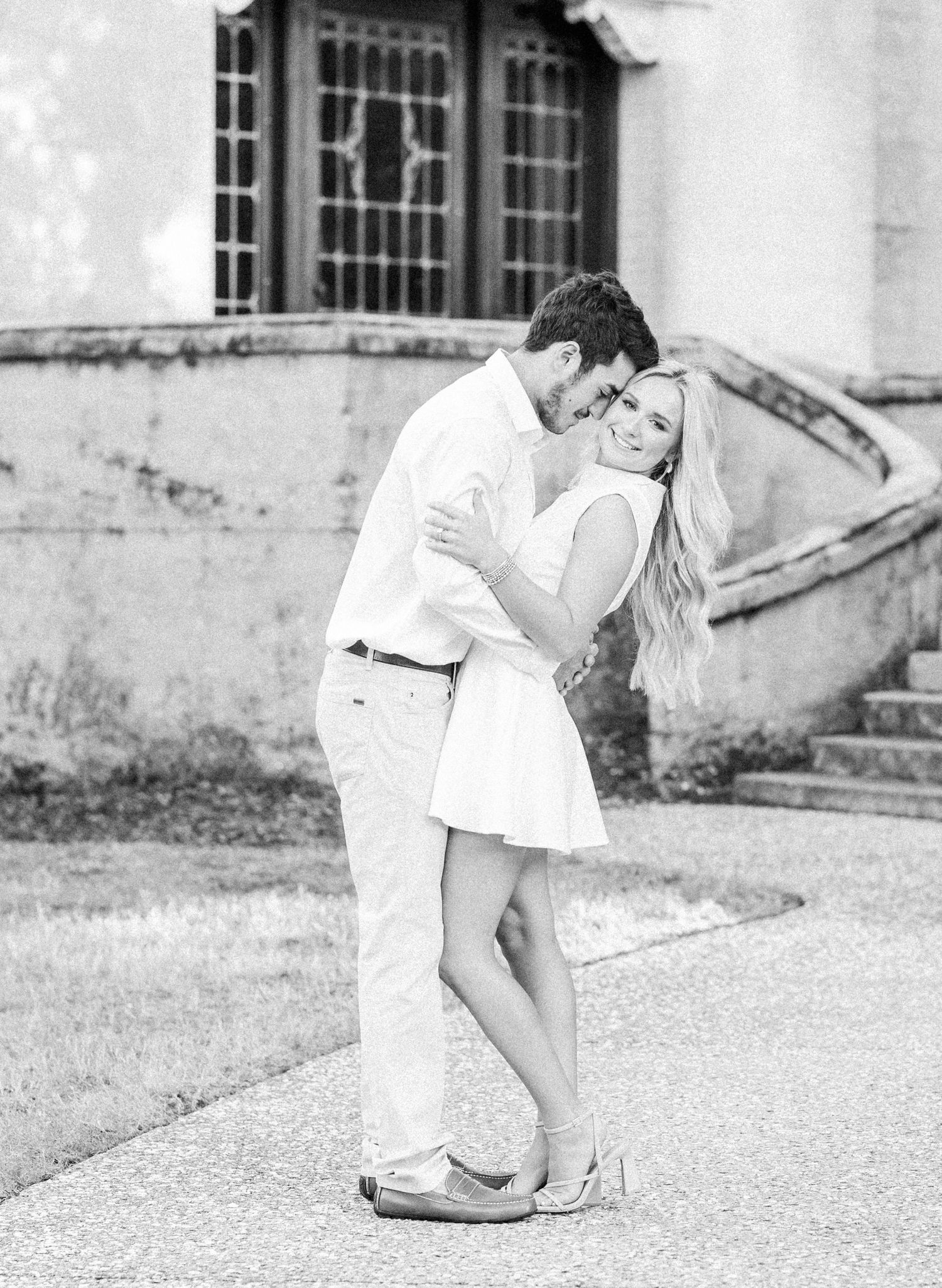 A romantic black and white portrait of a couple embracing outdoors on a paved walkway near a vintage building.