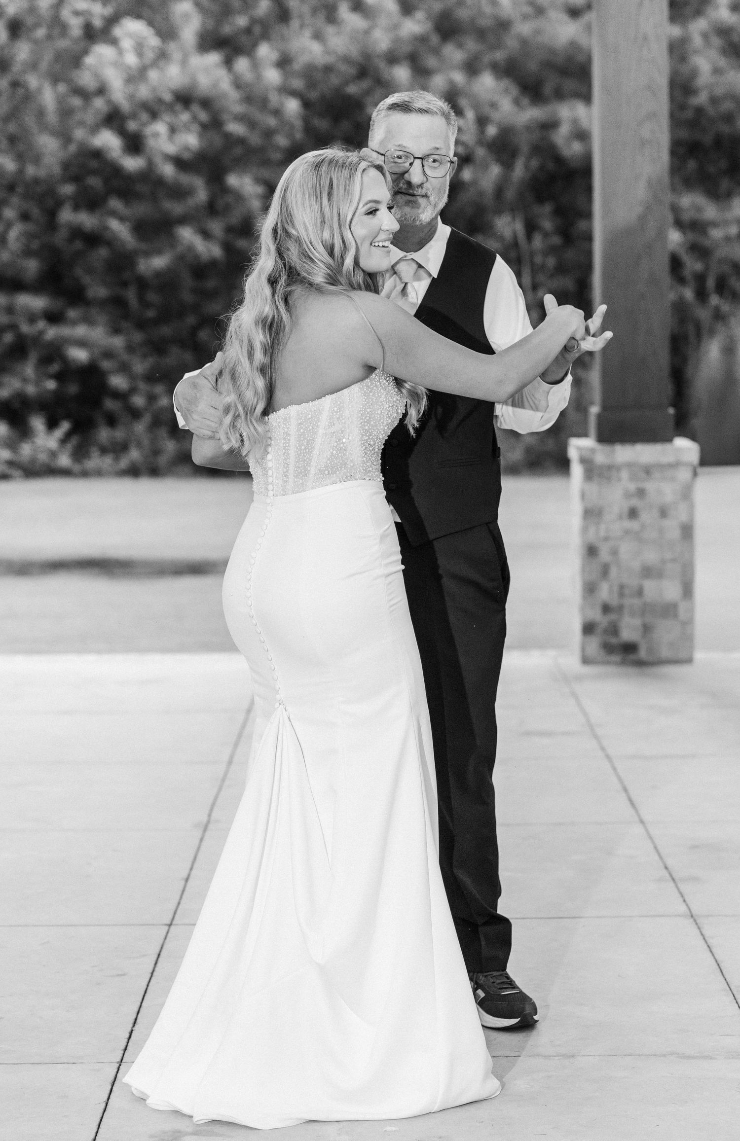 Black and white photo of bride in fitted gown dancing with father at outdoor wedding reception.