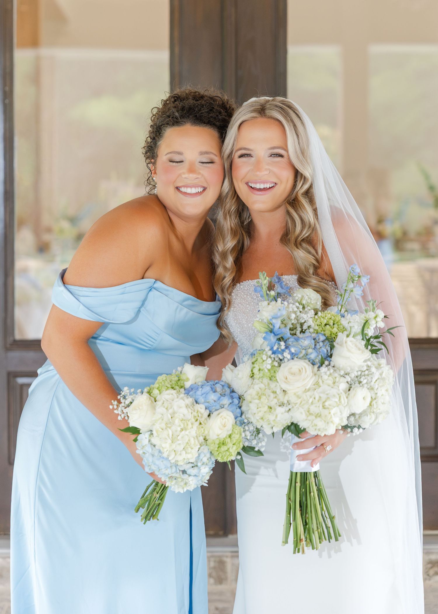 Bride in lace gown and bridesmaid in blue off-shoulder dress smiling together holding white floral bouquets.