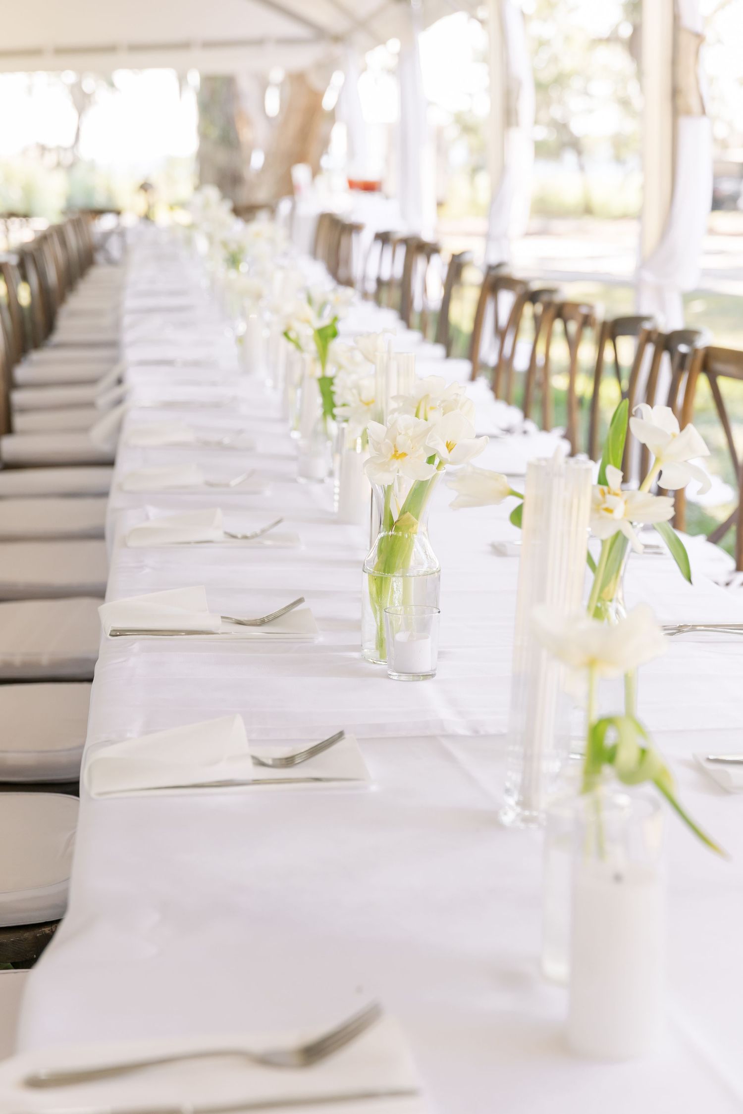 White linen covered banquet tables with delicate floral arrangements and place settings in an outdoor tent setting.