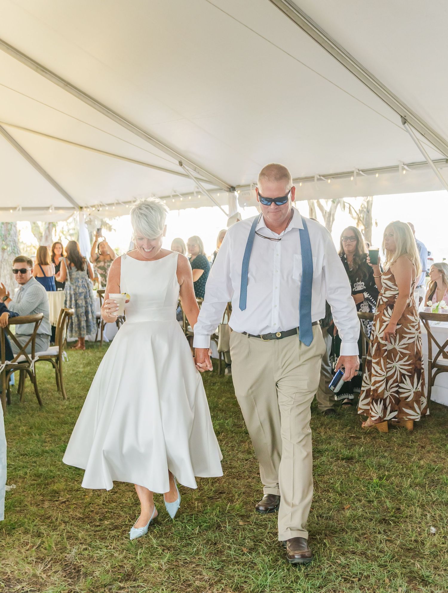 Wedding guests in casual summer attire dance and mingle under a white outdoor tent on grassy ground.
