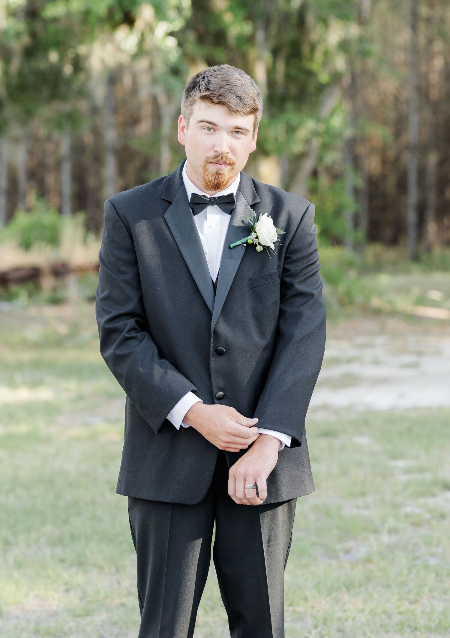 Handsome groom in black tuxedo with bow tie and white rose boutonniere posing outdoors at wedding venue.