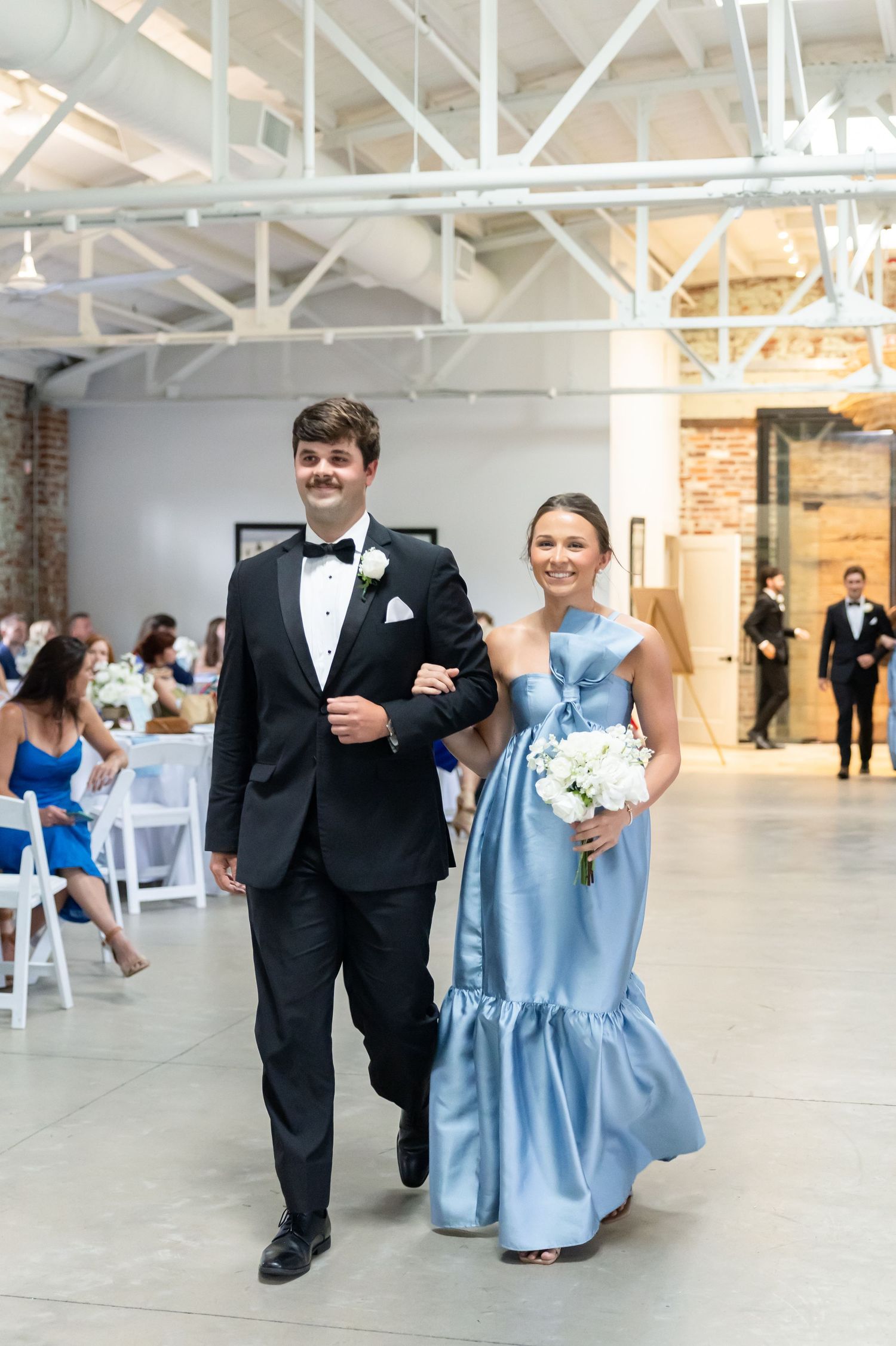 Groomsman escorts bridesmaid in blue gown down aisle at elegant indoor wedding ceremony.