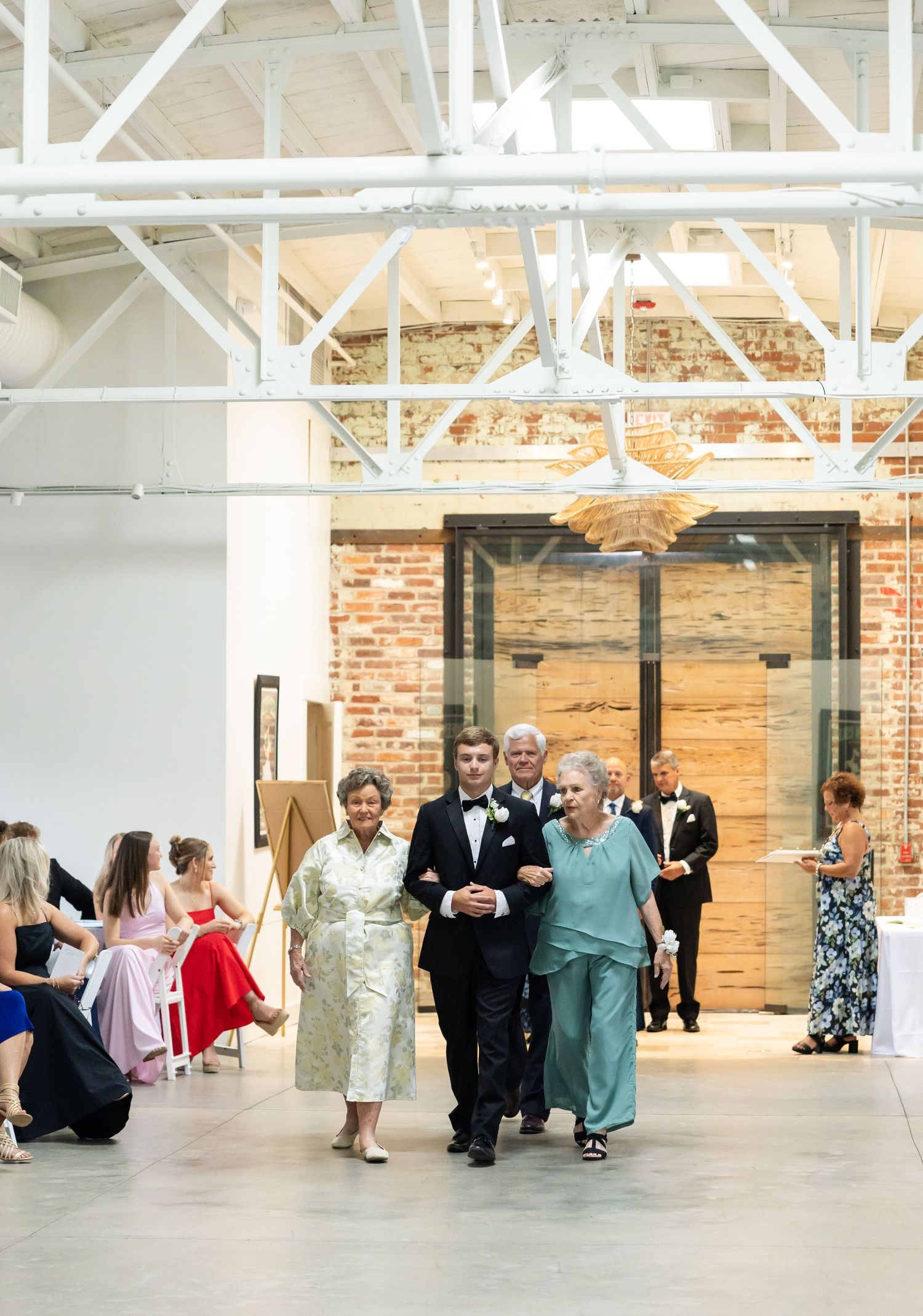 Groom escorted down aisle by two women in formal attire at elegant industrial wedding venue.