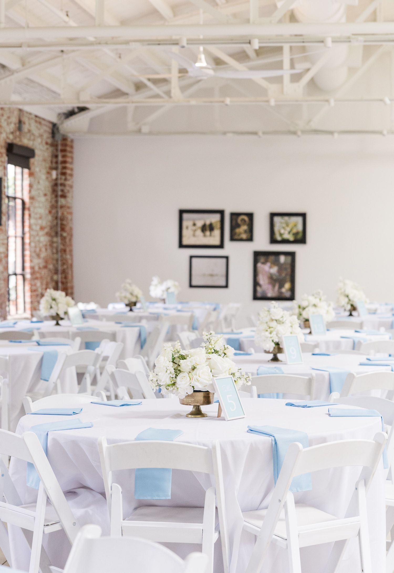 Elegant wedding reception hall with white round tables, light blue napkins, floral centerpieces, and white folding chairs.