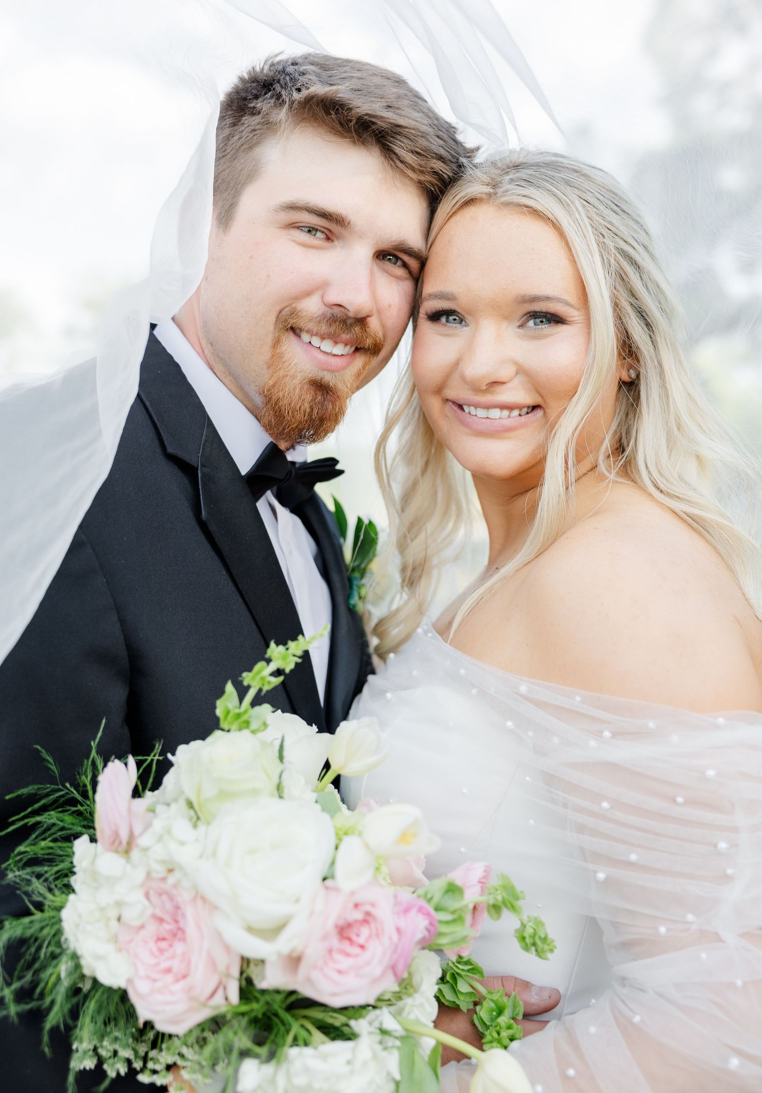Bride and groom smiling under a veil, bride holding a pink and white bouquet in a pearl off-shoulder gown.