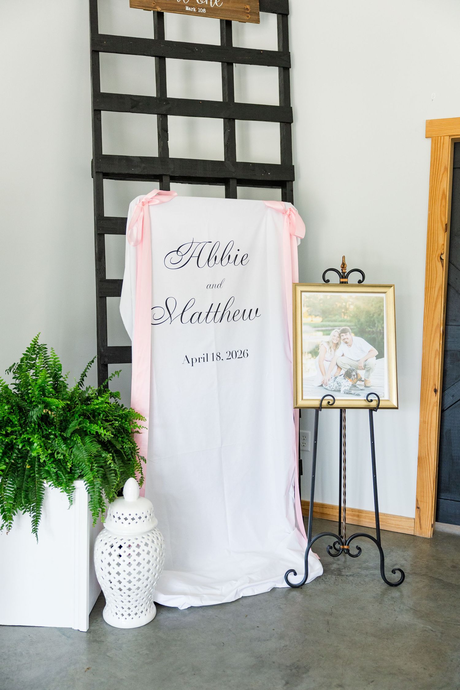 Elegant wedding welcome sign for Abbie and Matthew, April 18, 2026, with pink ribbon and framed photo on easel.