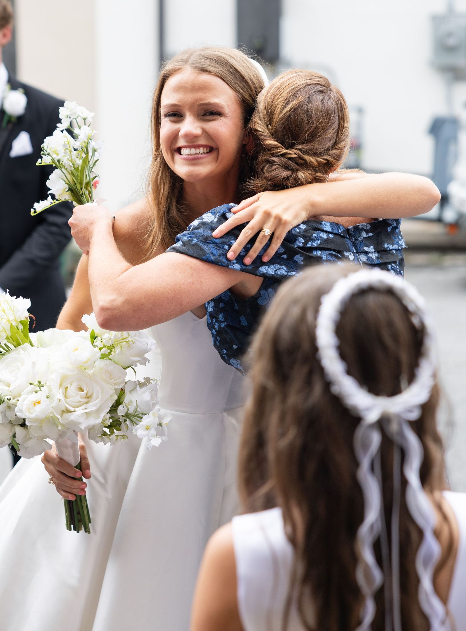 Smiling bride in white gown embraces guest while holding white floral bouquet after ceremony.
