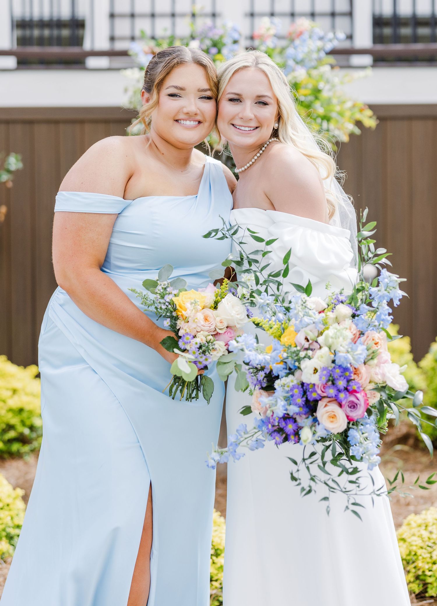 Bride in off-shoulder white gown posing with bridesmaid in light blue dress holding bouquets.
