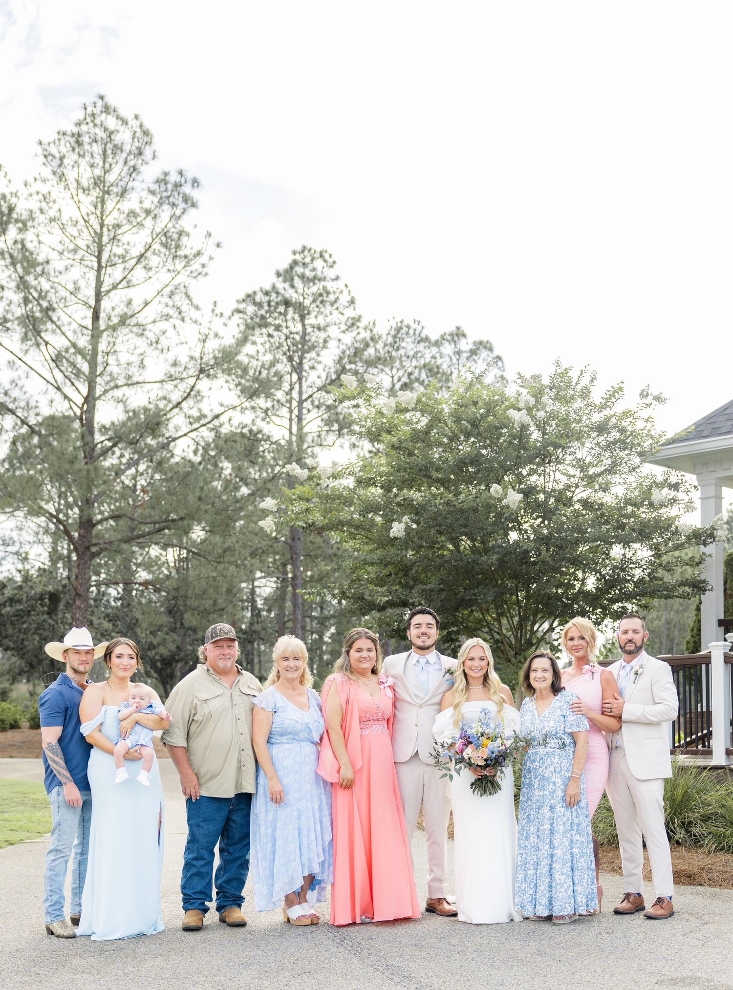 Wedding party group of ten people pose together outdoors in formal attire among tall pine trees.