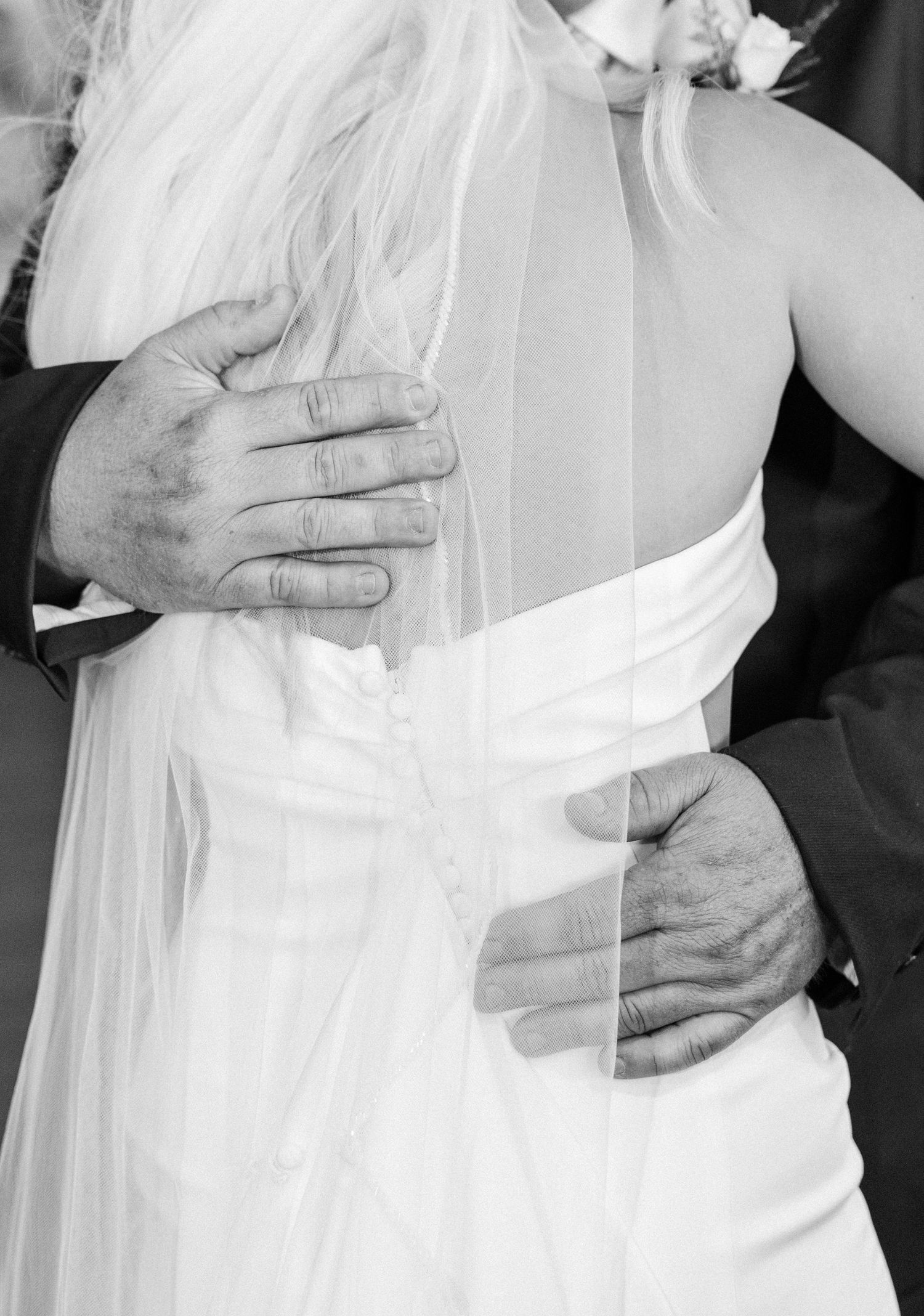 Black and white wedding photo of bride in white gown and veil being embraced from behind by groom.