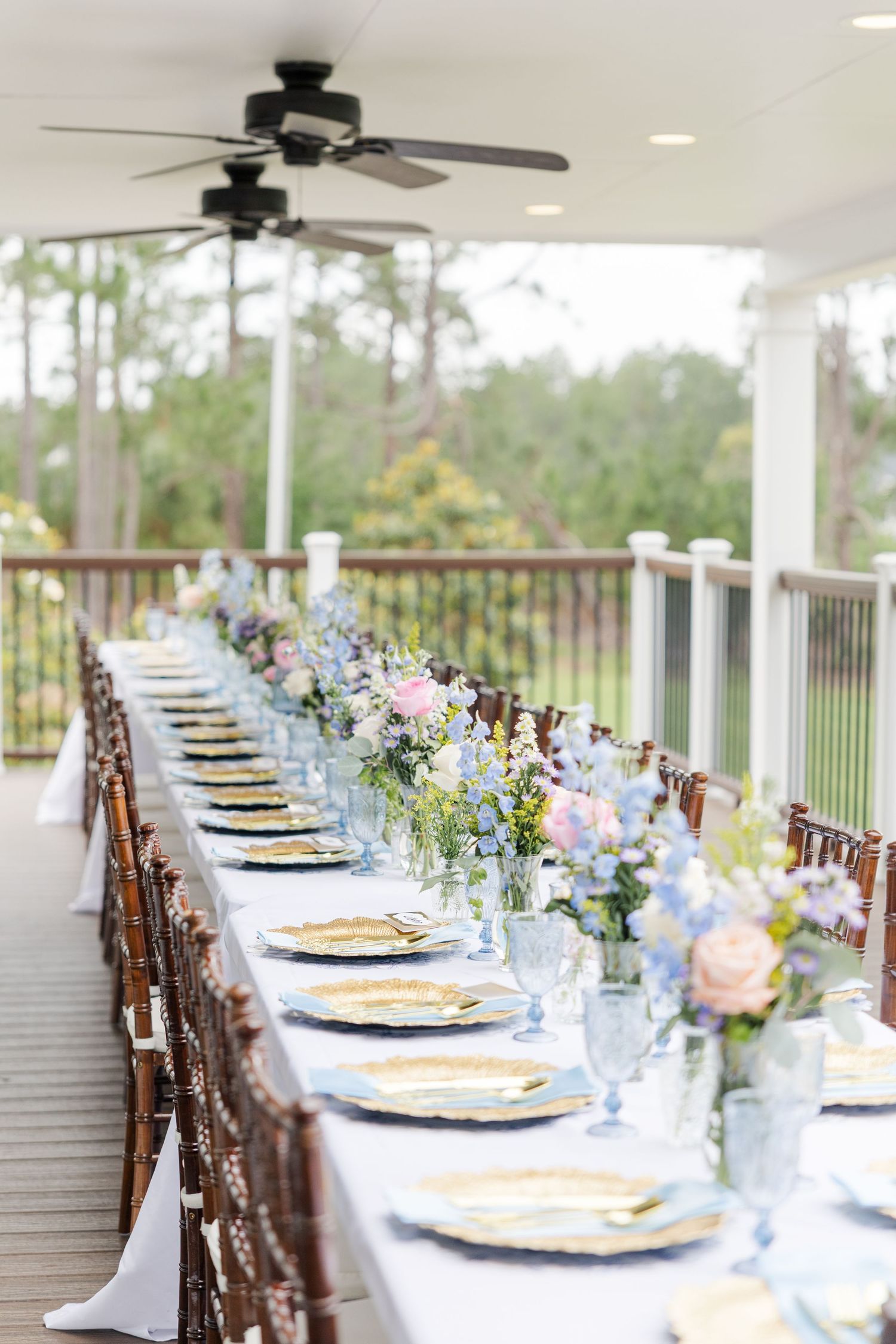 Elegant outdoor wedding reception table set with blue glassware, gold chargers, and pastel floral centerpieces.