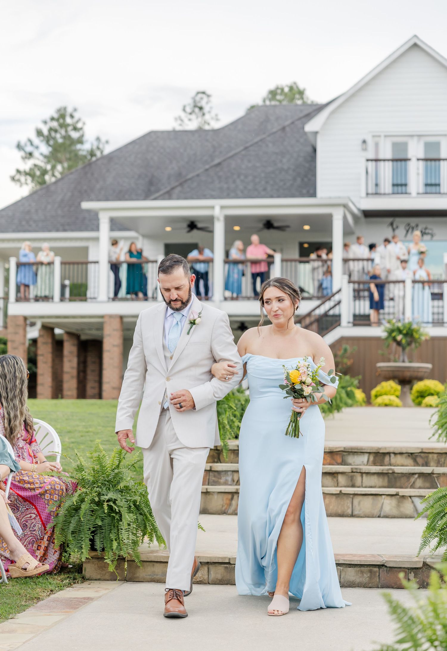 A groomsman escorts a bridesmaid in a light blue gown down the aisle at an outdoor wedding.