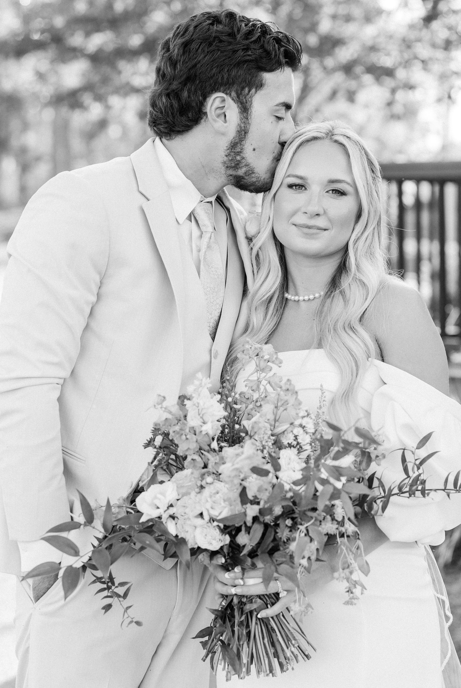 Black and white portrait of groom kissing bride on temple while she holds lush floral bouquet.