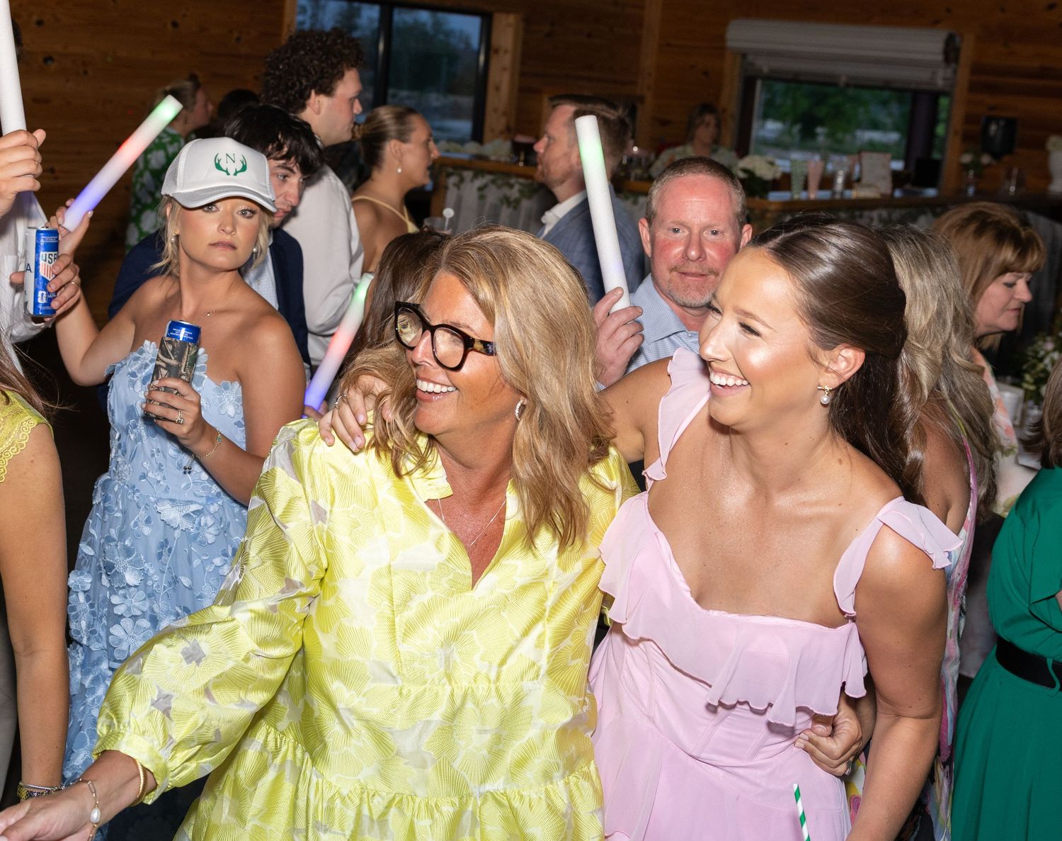 Guests celebrate at wedding reception holding glow sticks, woman in yellow dress dancing joyfully.