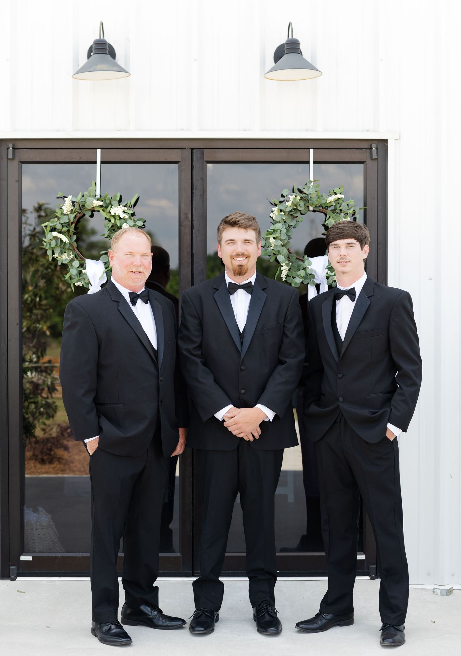 Three men in black tuxedos pose formally before white barn doors decorated with greenery wreaths.