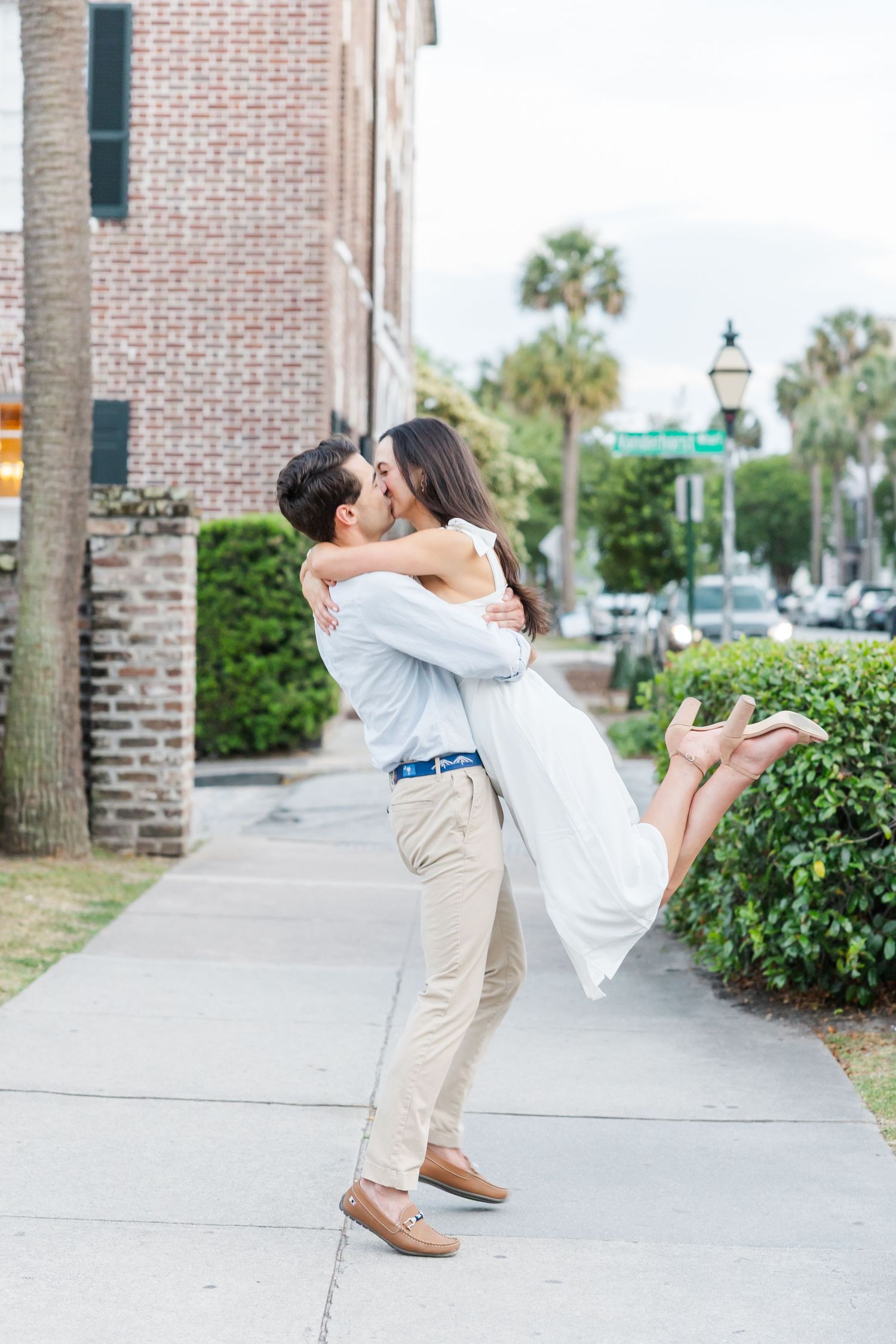 Man lifts and kisses woman on a city sidewalk, she wears a white dress, palm trees and brick building in background.
