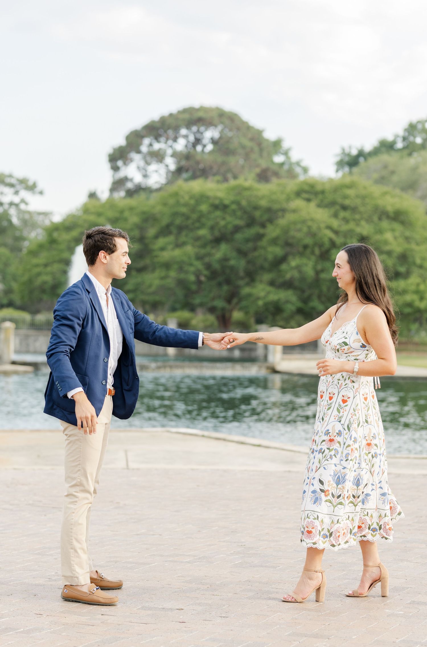 A couple holding hands and smiling at each other near a serene fountain in an outdoor park.